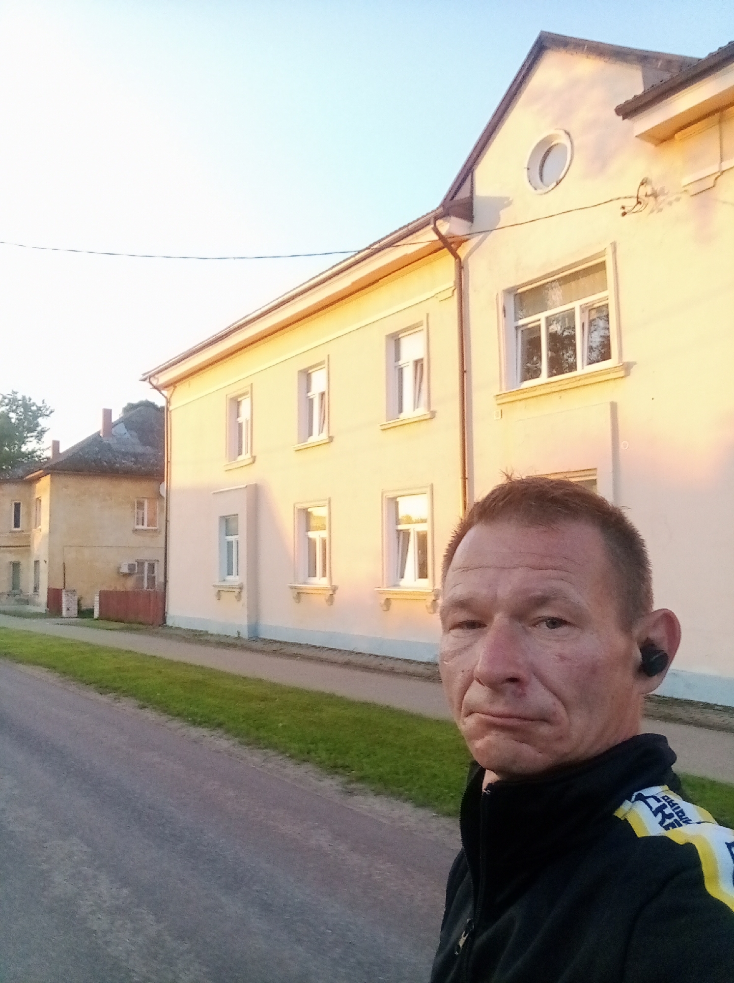 The image shows a man with short red hair, wearing a black jacket with yellow and white stripes on the sleeves, and an earpiece in his right ear. He is looking at the camera with a neutral expression. He is standing on a paved road, with a grassy verge to his left. Behind him are two buildings, likely residential, with cream-colored facades and numerous windows. The building on the right is closer and larger, with a prominent gabled roof. The building on the left is further away and appears older, with a darker, textured roof. The sky is a pale blue, suggesting it is either early morning or late afternoon. The scene appears to be in a quiet, possibly rural or suburban area. The style of the buildings and the overall atmosphere are consistent with locations in Tapa, Estonia, a city known for its railway junction and military base. The lighting on the buildings suggests sunlight at an angle, casting shadows and highlighting the textures of the facades. The road appears to be a two-lane street with no visible traffic in the immediate vicinity.