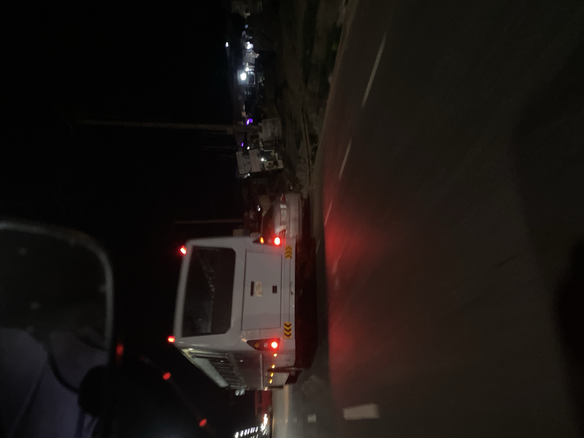 The image shows a white bus with red taillights traveling on a dark road at night in what appears to be Dam Camp, Nigeria. The bus is oriented to the right in the frame, with the back facing towards the camera. A dark vehicle's side mirror and part of the dashboard are visible on the left side of the frame, suggesting the image was taken from inside another car. The right side of the image shows the edge of the road and some dark vegetation. Streetlights are visible in the background.