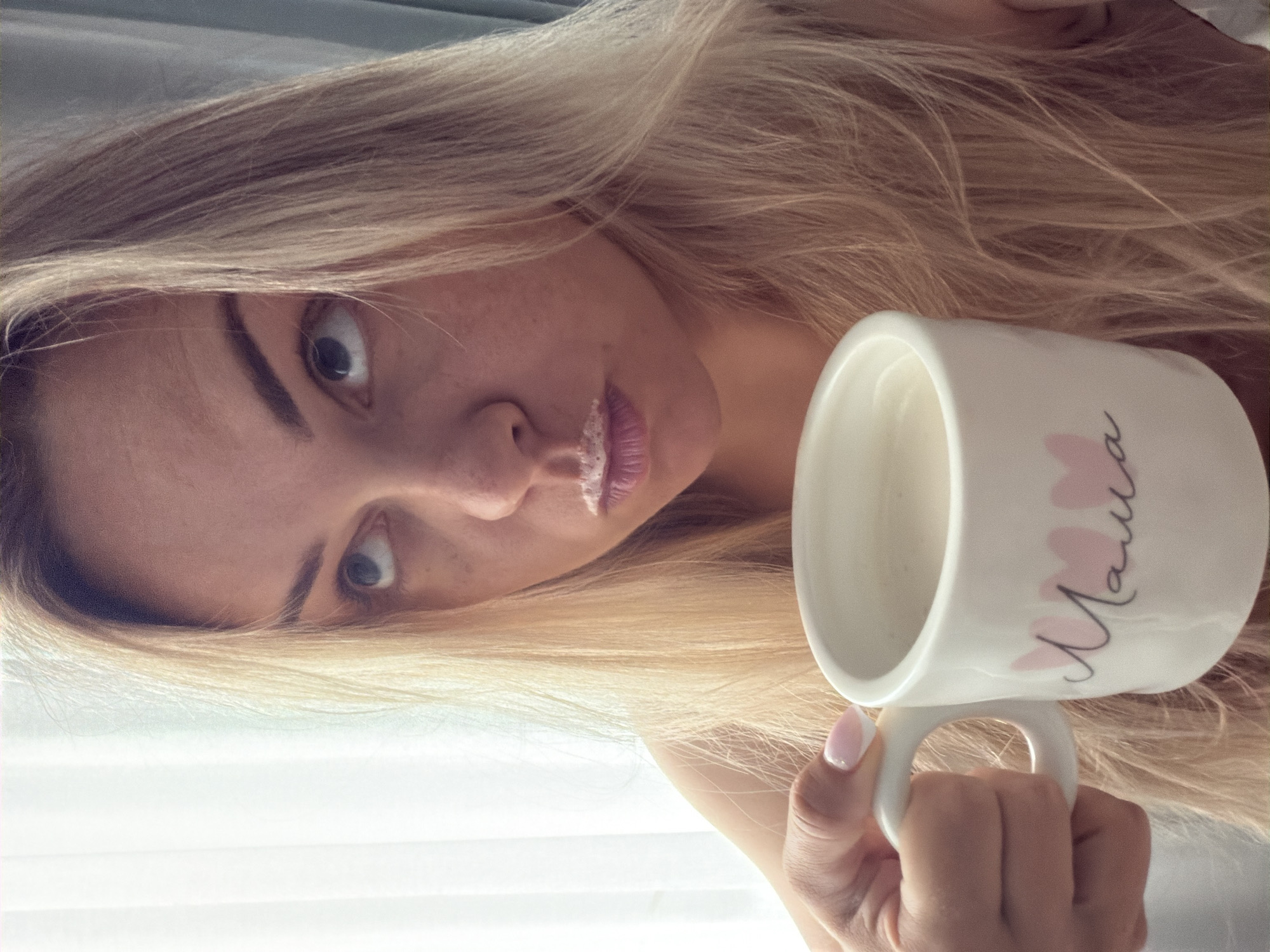 A fair-skinned woman with long blonde hair is holding a white mug with the Russian word "Mama" printed on it in cursive with a pink flower design, seemingly having taken a sip from it, as indicated by the light-colored residue on her upper lip. Her gaze is directed off-camera. The background is a lightly lit, neutral-toned interior. There is no discernible evidence to establish that the photo was taken in Votkinsk, Russia.