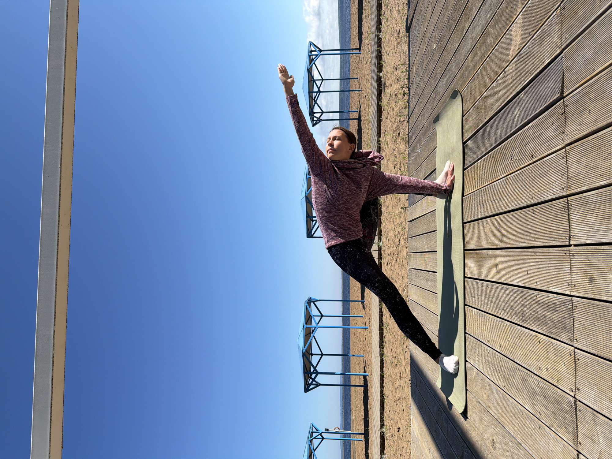 A young woman is performing a yoga pose on a wooden deck by a beach. She is wearing a long-sleeved hooded shirt and patterned leggings. Her left hand is on the yoga mat, and her right arm is extended upwards. The woman is positioned in a side plank variation or similar pose. The background features a sandy beach with several blue gazebos, a body of water, and a clear blue sky. The scene is set on a sunny day. The location is the beach in Chaikovsky, Russia.