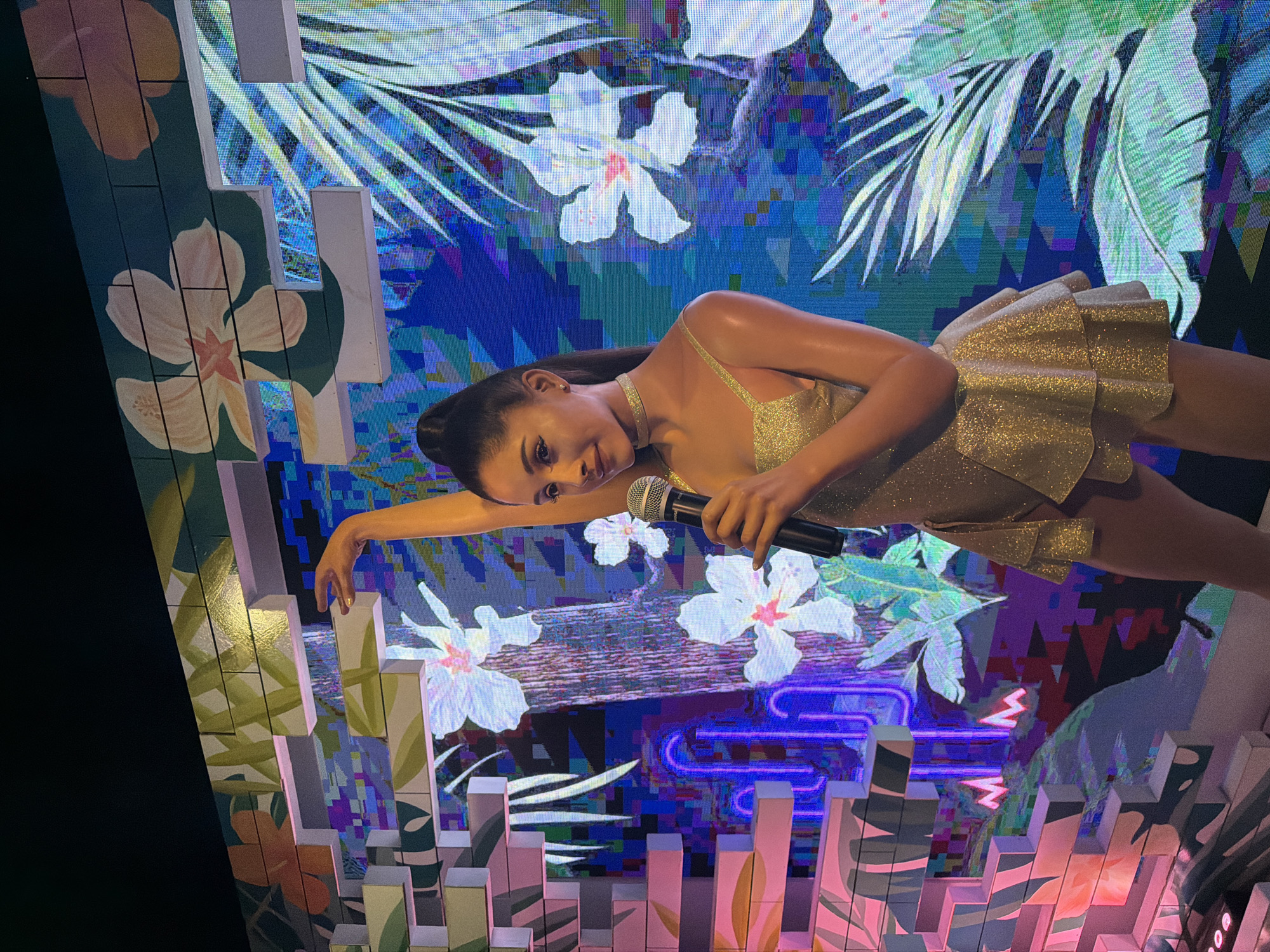 This is a photograph of a wax figure of Ariana Grande, a well-known singer, displayed in a tropical-themed setting at Madame Tussauds Bangkok, located in Siam Discovery, Bangkok, Thailand. The wax figure is wearing a sparkling gold dress with a tiered skirt and holding a microphone. The background features a vibrant digital display with images of tropical flowers, palm leaves, and neon lights, creating a lively and colorful environment. The lighting is bright and colorful, highlighting the wax figure and the detailed backdrop. The composition of the image focuses on the wax figure, with the background providing context and atmosphere.