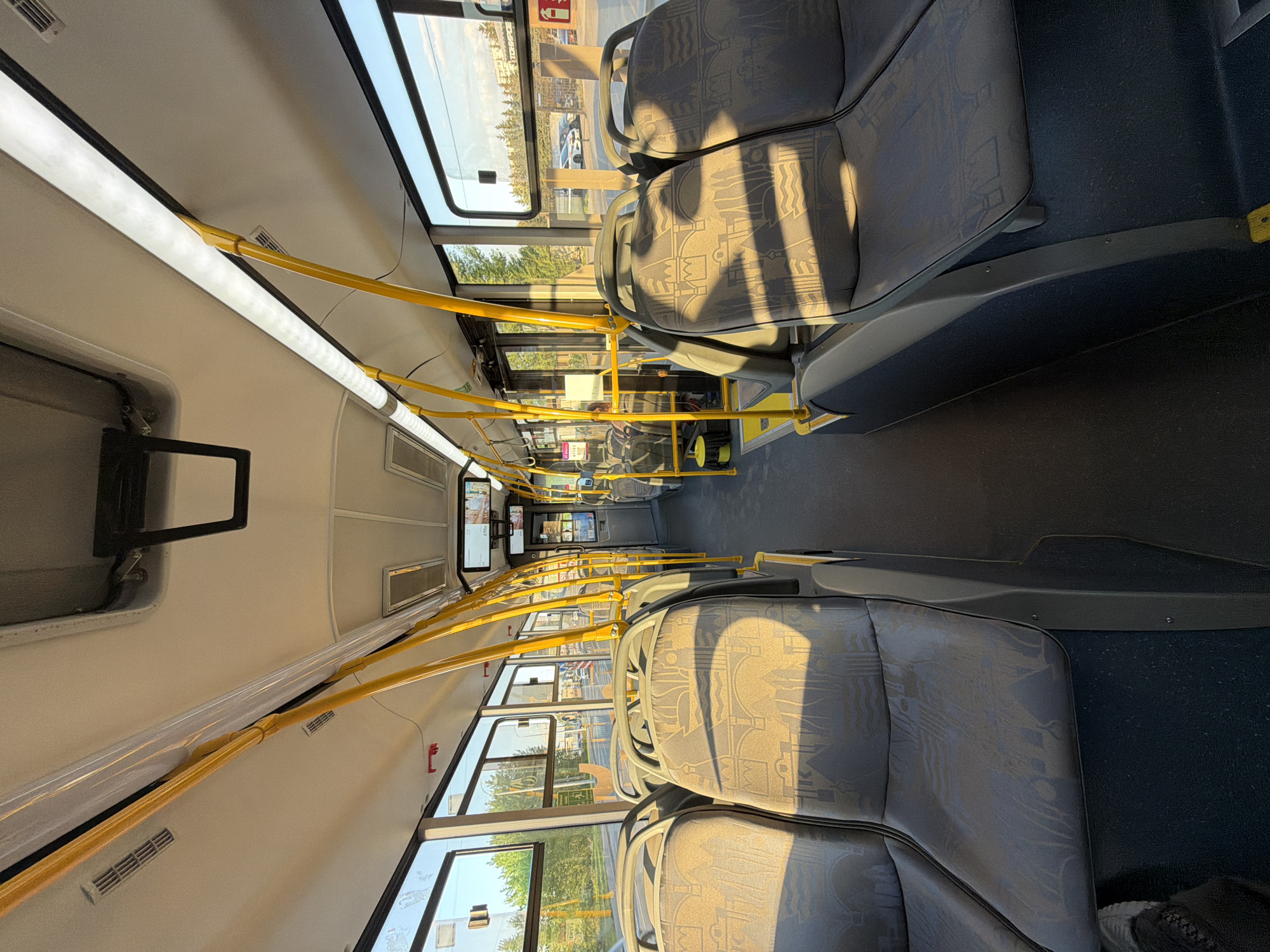 The image shows the interior of a bus, likely in transit. The seating is arranged in rows along the sides of the aisle. The seats are patterned in a gray and beige color scheme, with shadows cast across them, indicating sunlight entering through the windows. Yellow handrails run along the ceiling for standing passengers to hold onto. Windows line the sides of the bus, offering a view of trees and buildings outside, suggesting the bus is traveling in an urban or suburban area. The scene appears devoid of passengers except for a few visible towards the front of the bus. The lighting inside is bright, due to the combination of sunlight and overhead lights. The city of Krasnoyarsk, Russia is not definitively identified in the image.