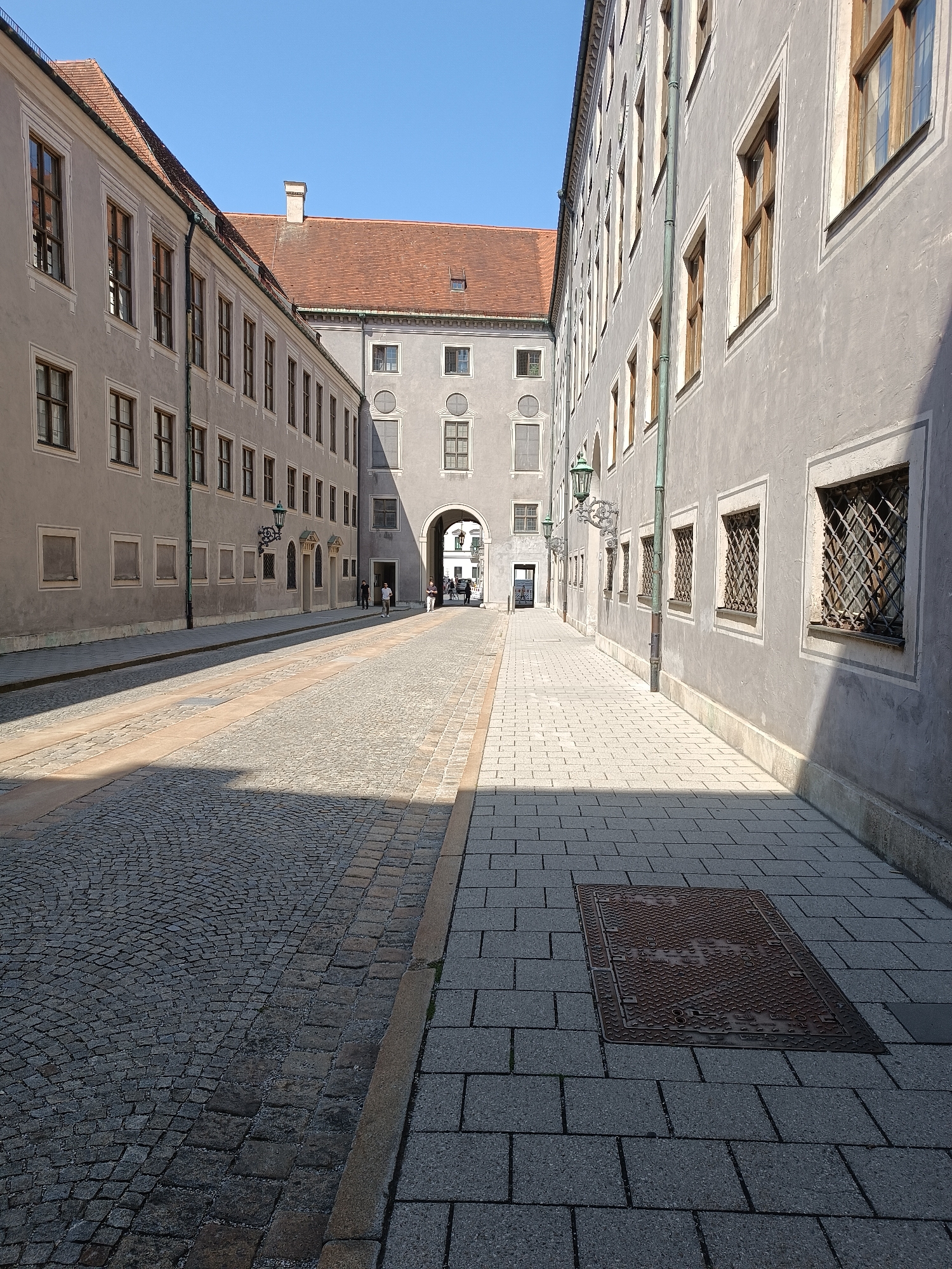 This image depicts a view down a cobblestone street in Munich, Germany, flanked by two imposing grey buildings. The street is divided into two sections by a slight elevation and a change in paving material. The left side features dark, rough cobblestones arranged in a semicircular pattern, while the right side is paved with lighter, rectangular concrete slabs. A manhole cover with a decorative, textured surface is visible on the right-hand pavement.
The buildings on either side are multi-story structures with numerous windows. The building on the left has a reddish-tiled roof and appears to be part of a larger complex. The building on the right has a simpler, flat roof and is characterized by its rows of windows, some of which are barred. The architecture suggests a historical or institutional setting, possibly a palace or a museum.
In the distance, an arched gateway is visible, leading to another courtyard or street. A few individuals are walking in the distance, and a horse-drawn carriage is partially visible through the arch. The sky is a clear, bright blue, indicating a sunny day, and long shadows are cast across the street by the buildings. The overall impression is one of a quiet, sun-drenched urban pathway within a significant historical district of Munich.