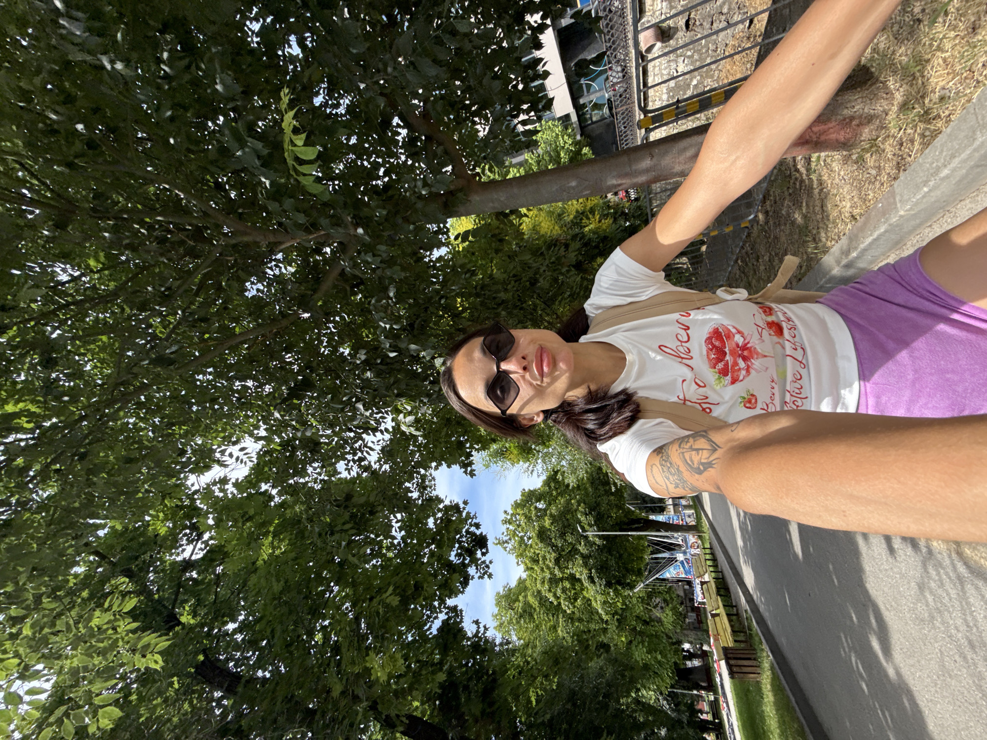 The media file shows a woman taking a selfie outdoors, surrounded by lush green trees. The camera is positioned low, looking upwards towards the subject and the canopy of leaves. The woman is wearing sunglasses and a white t-shirt with a graphic of strawberries and text. She also has on purple shorts. Her right arm is visible, showing a tattoo. The scene appears to be a park or a tree-lined street, with a paved path and a metal fence visible in the background. Sunlight filters through the leaves, suggesting it is daytime, likely morning or afternoon. The overall mood seems casual and relaxed, with the woman appearing to enjoy a sunny day outdoors. There is some graffiti on a building in the background. The text on her shirt reads "Berry Lifesyle".