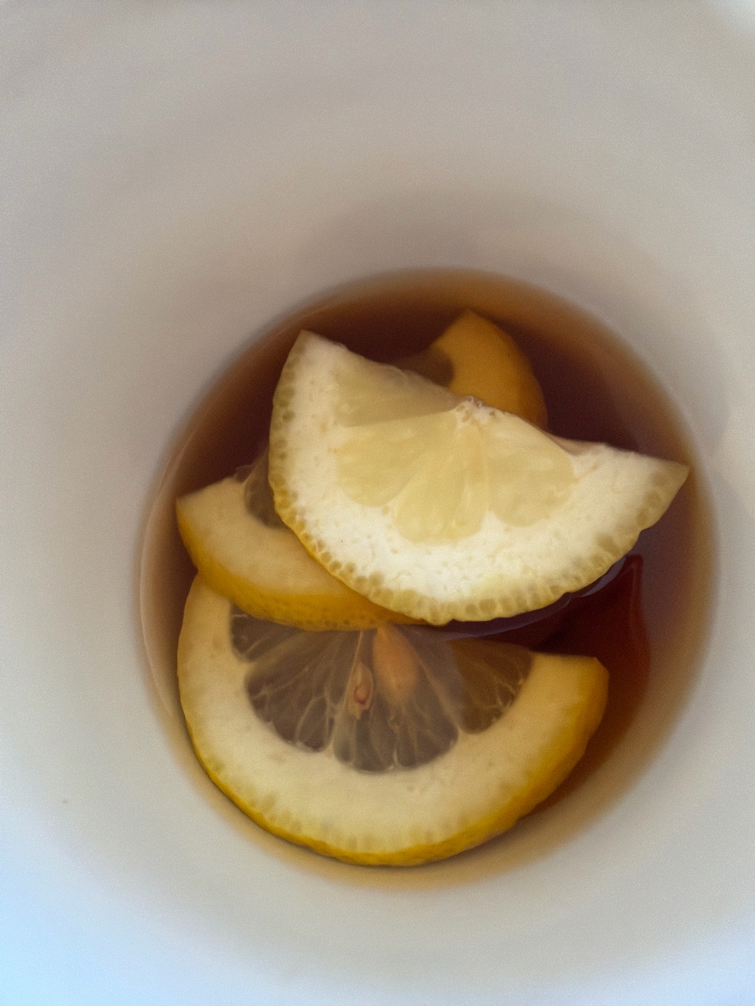 A close-up, top-down shot reveals three lemon slices submerged in a dark liquid within a white mug. The lemon slices, each with visible seeds and pulp, vary in their placement and overlap. The liquid is a warm, amber hue, suggesting it could be tea or a similar beverage. The mug's interior is clean and smooth, with the focus solely on the lemons and the liquid. The background is predominantly the white interior of the mug. There are no people or other objects visible in the scene.