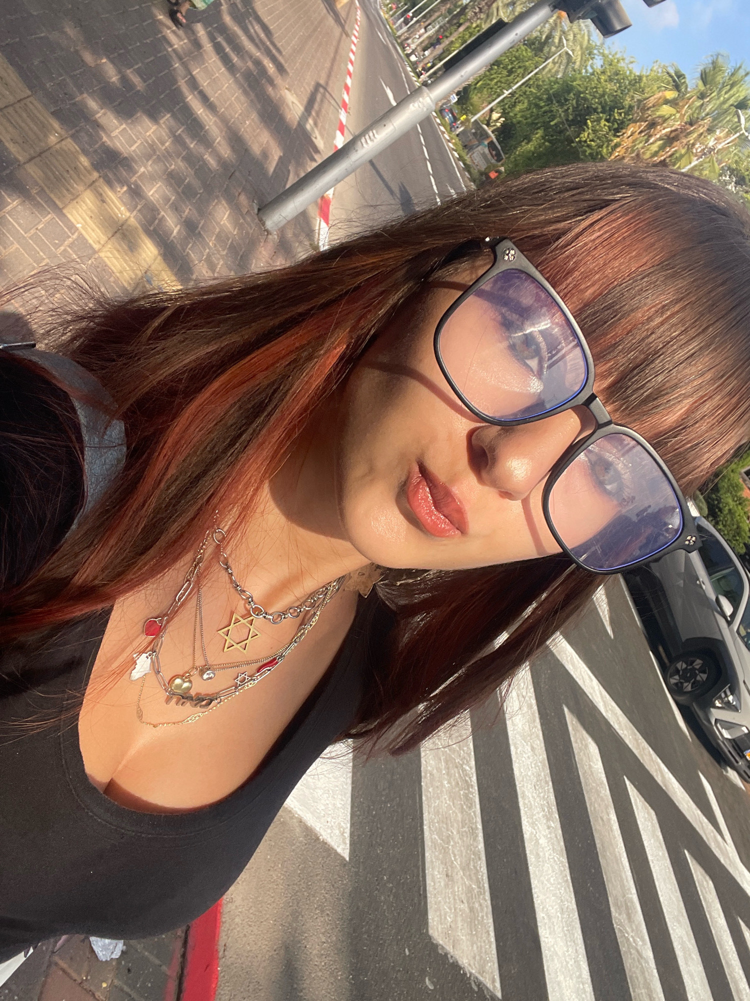 A young woman with auburn hair and a black tank top is taking a selfie outdoors. She is wearing glasses and multiple layered necklaces, including one with a Star of David pendant. She is standing on a pedestrian crossing with white stripes. In the background, there is a street with palm trees and a car. The scene appears to be in the city of Raanana, Israel, as suggested by the presence of palm trees and the general urban environment. The lighting suggests it is daytime, likely late afternoon given the long shadows.