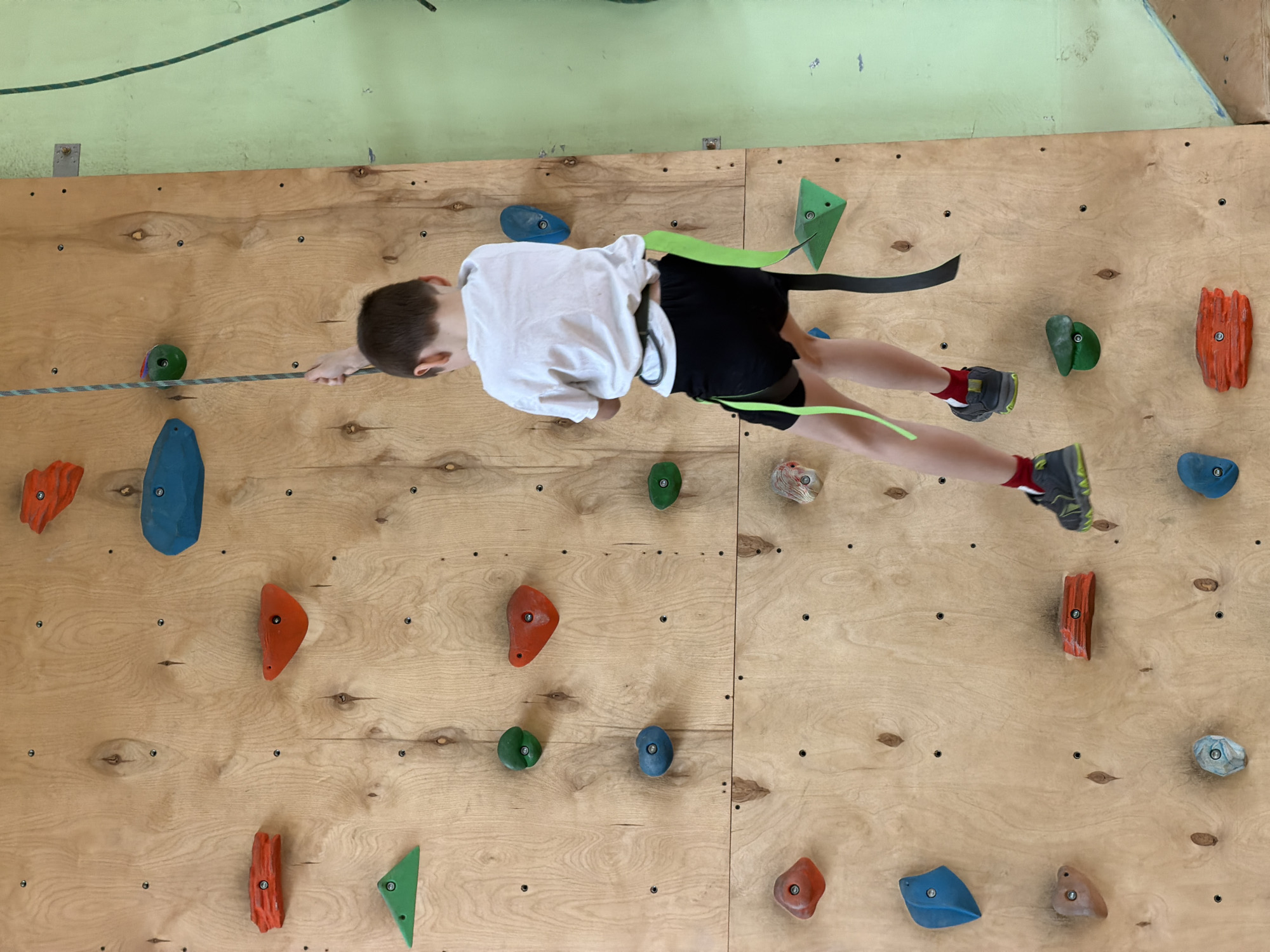 A young boy is captured in action, rock climbing on an indoor wall. He is positioned horizontally across the wooden surface, supported by a climbing harness with black and neon green straps, and secured by a grey and white rope that runs across the wall. The boy is wearing a white short-sleeved t-shirt, black shorts, and grey and lime green climbing shoes with red socks.
His left hand is extended towards a green climbing hold, while his right hand holds onto the rope near his body. His legs are bent, with his feet planted on lower climbing holds. The climbing wall is made of light-colored plywood panels, onto which numerous colorful climbing holds of various shapes and sizes are bolted. These holds are predominantly orange, blue, and green, with some lighter-colored ones as well. Above the wooden panels, the wall is painted a faded light green. The overall setting is an indoor climbing facility, likely a gym or training area, potentially in Izhevsk, Russia. No visible text is present in the image.