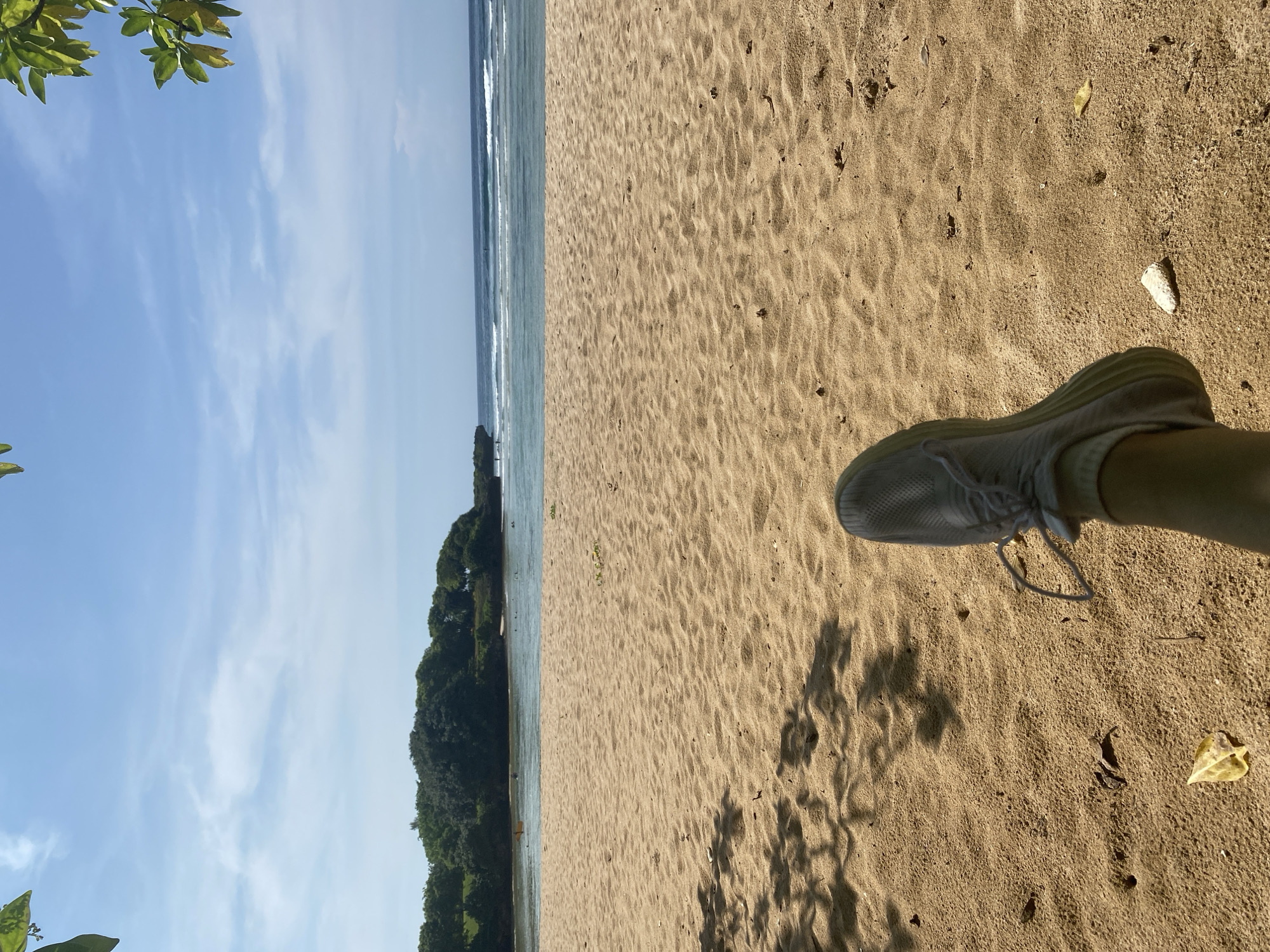 The image depicts a sandy beach scene in Nusa Dua, Indonesia. The perspective is from a low angle, showing a foot with a white sneaker in the foreground, casting a shadow on the sand. The sand has various textures, including footprints and small debris. The background features the ocean with gentle waves and a line of trees/vegetation separating the sand from the water. A clear blue sky with scattered clouds is visible above.
