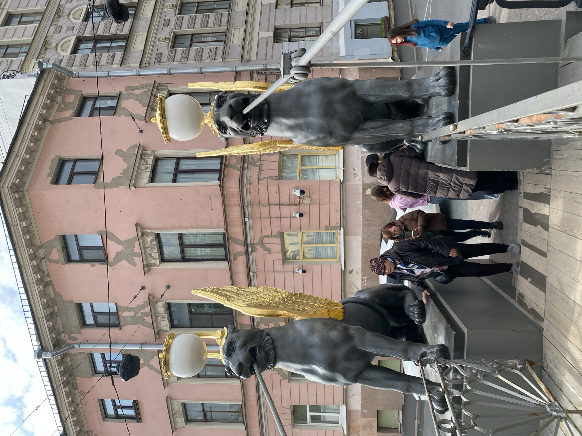 The image depicts the Bank Bridge in Saint Petersburg, Russia. Two winged lion statues are prominent in the foreground. They are grey with golden wings and each holds a white globe-shaped lamp in its mouth. Several people are present on the bridge. Some are looking at the statues. The background features a pink building with windows. The sky is visible in the upper left corner.