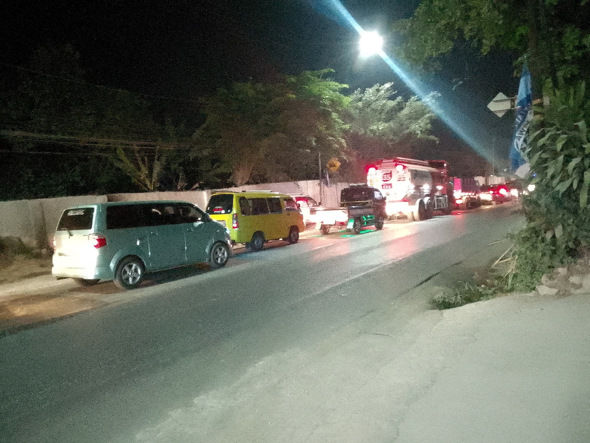 This is a night-time, ground-level, eye-level shot of traffic on a paved road in Gunungmasigit, Indonesia. The road is mostly empty, with a few vehicles parked on the side and a small line of traffic in the distance. The sky is dark, with a bright street light illuminating the scene from the top left. Trees and foliage are visible on both sides of the road. The vehicles visible include a light blue minivan, a yellow minivan, a truck, and a motorcycle. The blue minivan is parked on the left side of the road. The yellow minivan is behind it, also parked. Further down the road, a truck is visible, along with other vehicles in the distance. A blue flag is attached to a pole on the right side of the road, near some bushes. The overall scene suggests a quiet, rural road at night.