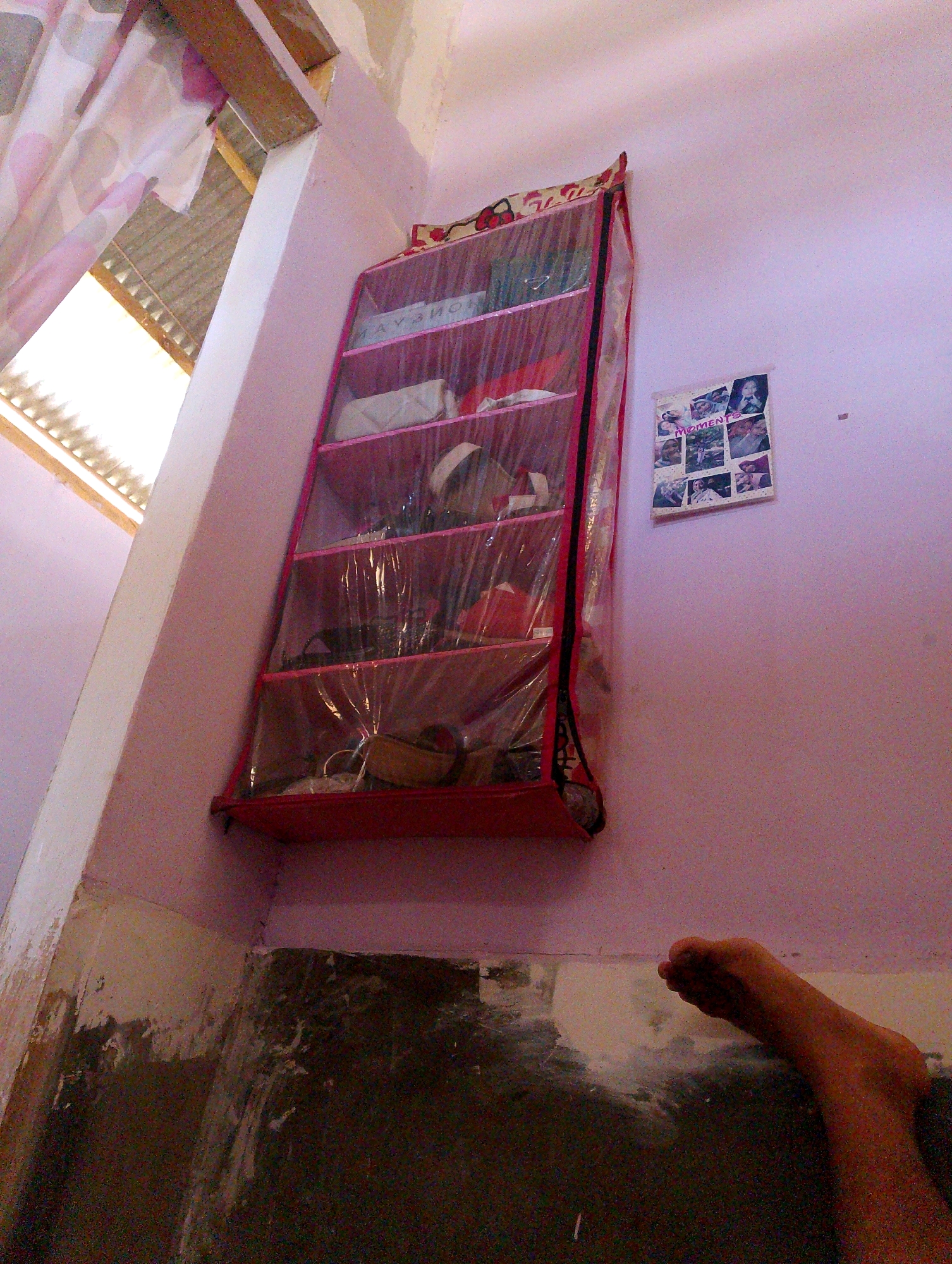 The image depicts an interior scene, likely a room in a residential setting in Desa Pagandaran, Indonesia. The primary focus is a multi-tiered shoe organizer made of pink fabric and clear plastic, positioned against a pale purple wall. The organizer contains various items, though specific identification is difficult due to the plastic covering and lighting. A collage of framed photos with the word "MOMENTS" is hanging on the wall to the right of the organizer. In the foreground, a bare human foot is visible, resting on a dark, textured floor. The floor appears worn and partially covered with debris or discoloration. To the left of the organizer, a portion of a window with a patterned curtain and corrugated roofing is visible, suggesting the location might be close to an exterior wall or a verandah. The overall impression is that of a cluttered, lived-in space.