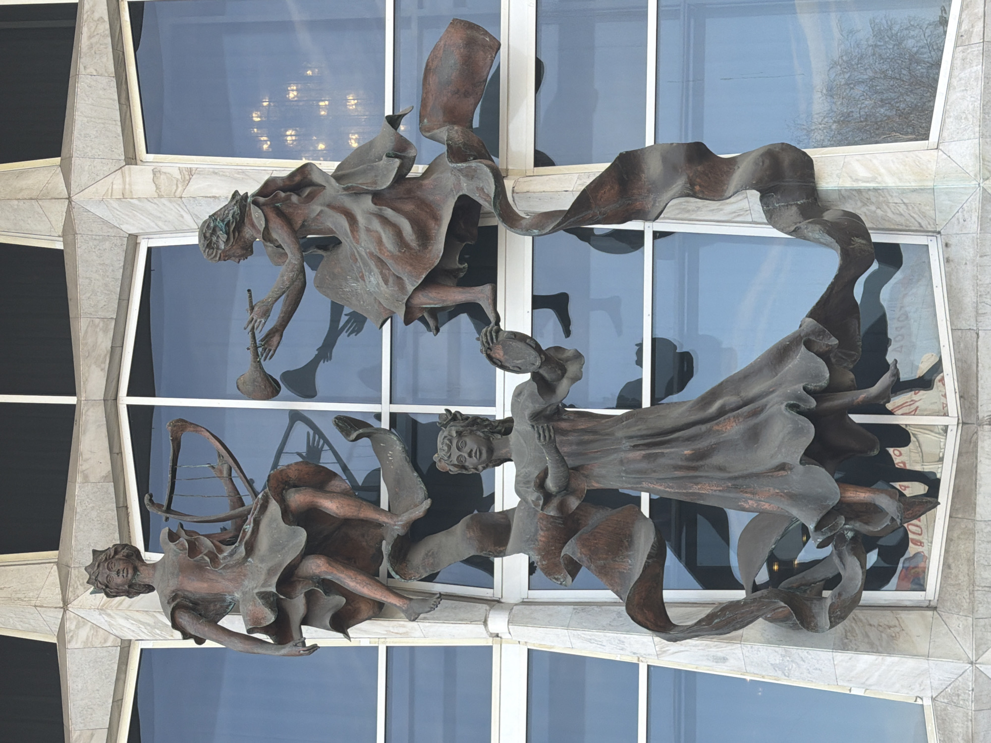 The image shows a bronze sculpture of three figures on the exterior of a building with large windows. The figures appear to be celestial beings or muses, draped in flowing garments. One figure holds a harp, another a horn or trumpet, and the third a small dish or offering. The sculpture is mounted on a building facade with marble or stone-like tiles and large reflective windows. The reflections in the windows suggest an interior with a chandelier and possible silhouettes of people. The location is the Krasnoyarsk Opera and Ballet Theater, Krasnoyarsk, Russia.