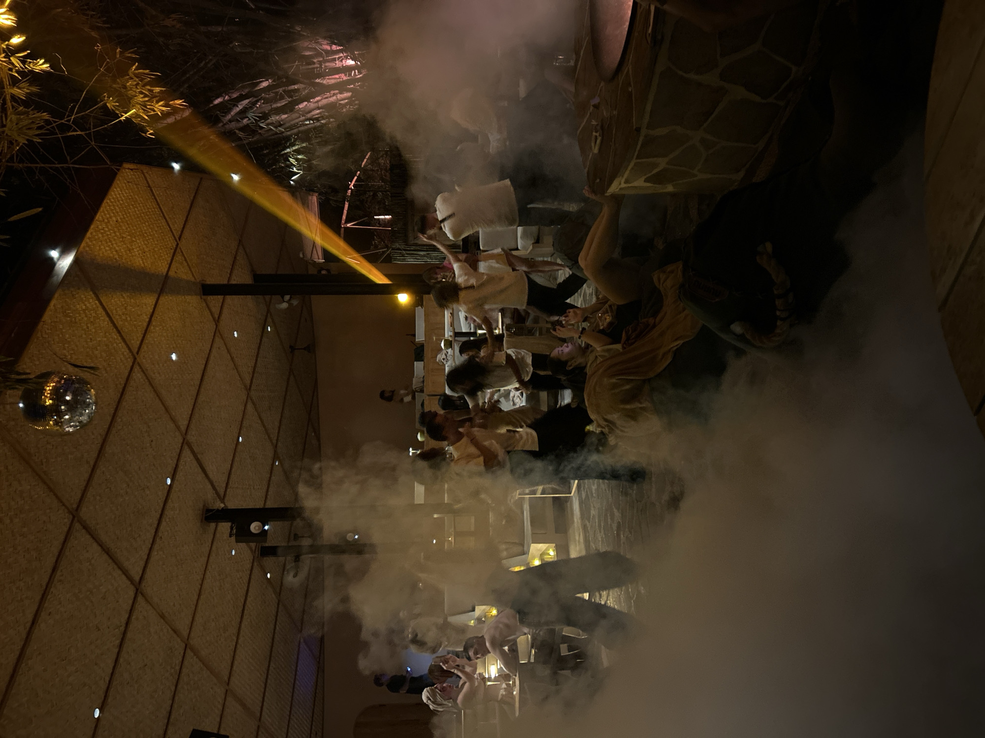 The image depicts a vibrant nighttime scene at a venue that appears to be an outdoor lounge or restaurant. The environment is characterized by a thatched roof, ambient lighting from spotlights and a disco ball, and a significant amount of fog or smoke creating a dramatic atmosphere.
In the foreground, several people are visible, some seated and others standing amidst the fog. One individual is lying down on the ground, partially obscured by the smoke, wearing light-colored clothing.
Further into the scene, a group of people are interacting, with some dancing or socializing. They are dressed in casual attire. The lighting includes a prominent orange beam of light cutting across the scene from the upper right.
In the background, there are tables and seating areas where other patrons are present. The overall impression is one of a lively social gathering, possibly a party or event. The venue's architecture suggests a tropical or bohemian aesthetic. The mention of Kediri, Indonesia, implies this is a local establishment.