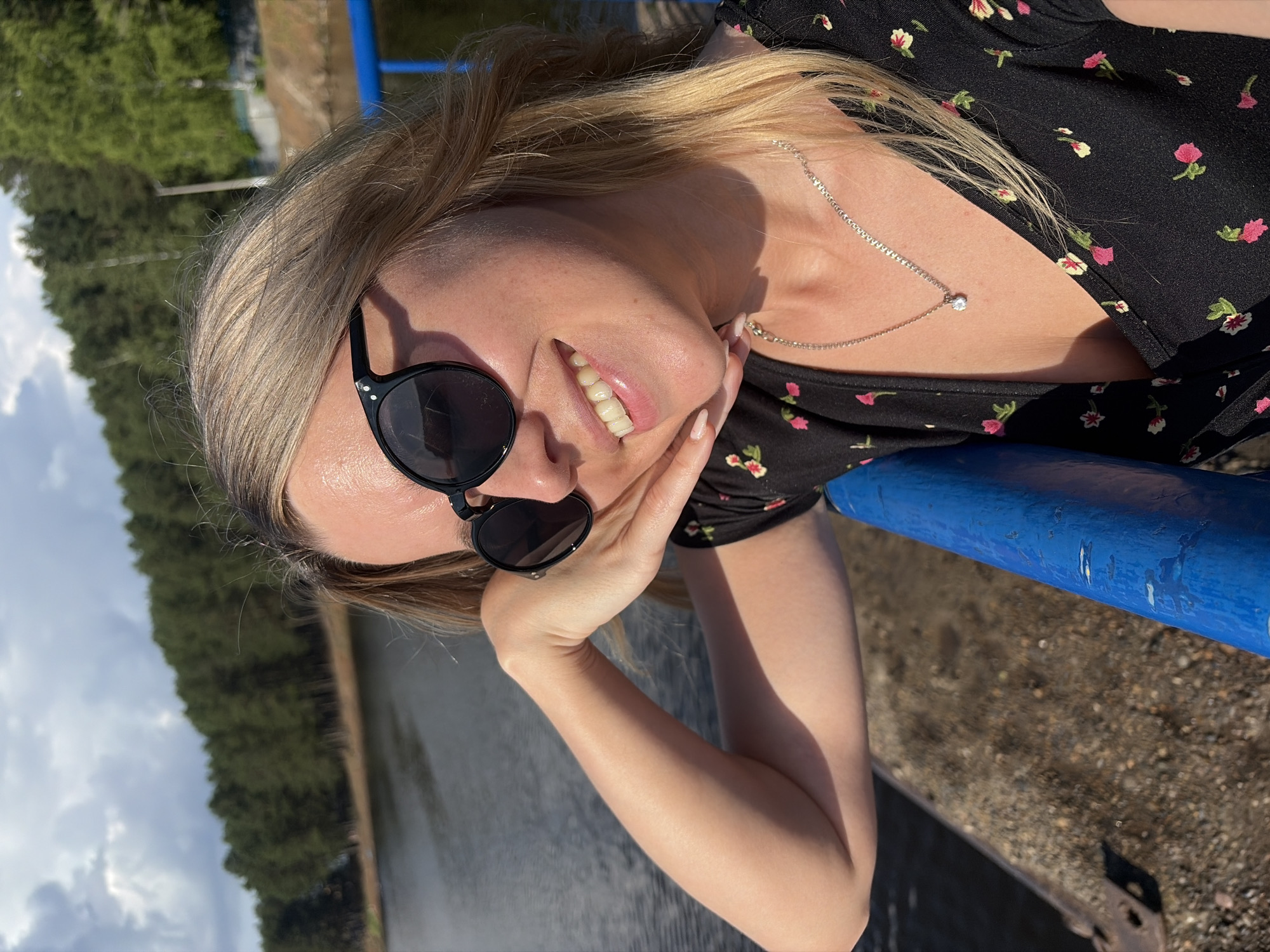 In a candid outdoor shot, a young Caucasian woman with blonde hair, wearing sunglasses and a black dress with a floral pattern, smiles warmly at the camera. She rests her chin on her hand, and a silver necklace with a small pendant adorns her neck. The background reveals a body of water, likely the Kama River, bordered by a dense forest of pine trees. A blue railing is visible in the foreground, suggesting she is on a pier or promenade in the city of Chaikovsky, Russia. The sky above is partly cloudy, with sunlight casting shadows.