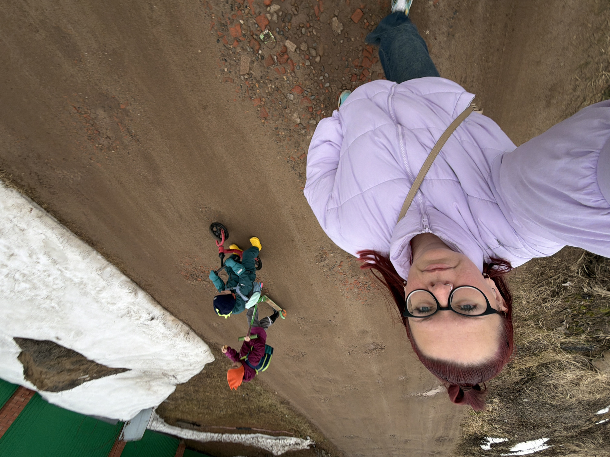 The image is an angled, downward selfie taken by a woman wearing glasses and a light purple puffer jacket. She has red hair pulled back in a bun and is looking at the camera. Below her are two children on a dirt road, one on a scooter and the other on a balance bike. In the bottom left corner is a patch of snow next to a green-roofed structure. The location is identified as Chaikovsky, Russia, based on the file properties.