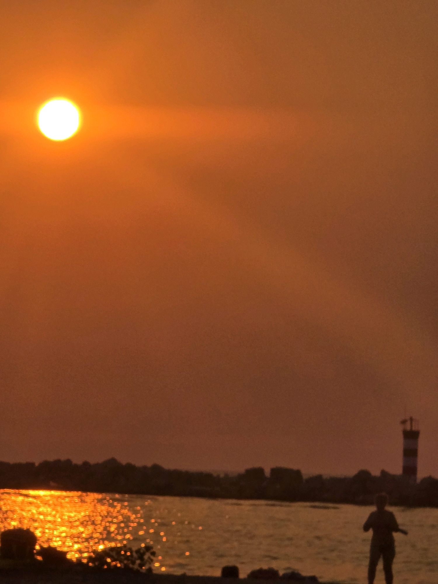 The image depicts a sunset over the sea, with the sun appearing as a bright, orange orb in the upper left quadrant. The sky is a gradient of orange and red hues. In the foreground, the water is illuminated by the setting sun, creating a shimmering effect with numerous points of light. A dark silhouette of a person is visible on the right side of the frame, standing near the water's edge and appearing to be facing the sea. Further in the background, a dark silhouette of a shoreline is visible, punctuated by what appears to be a lighthouse with red and white stripes. The overall scene suggests a coastal environment during sunset. Given the presence of a lighthouse and a coastal setting, this scene could plausibly be located in Bar, Montenegro, which has a coastline and a historical lighthouse.