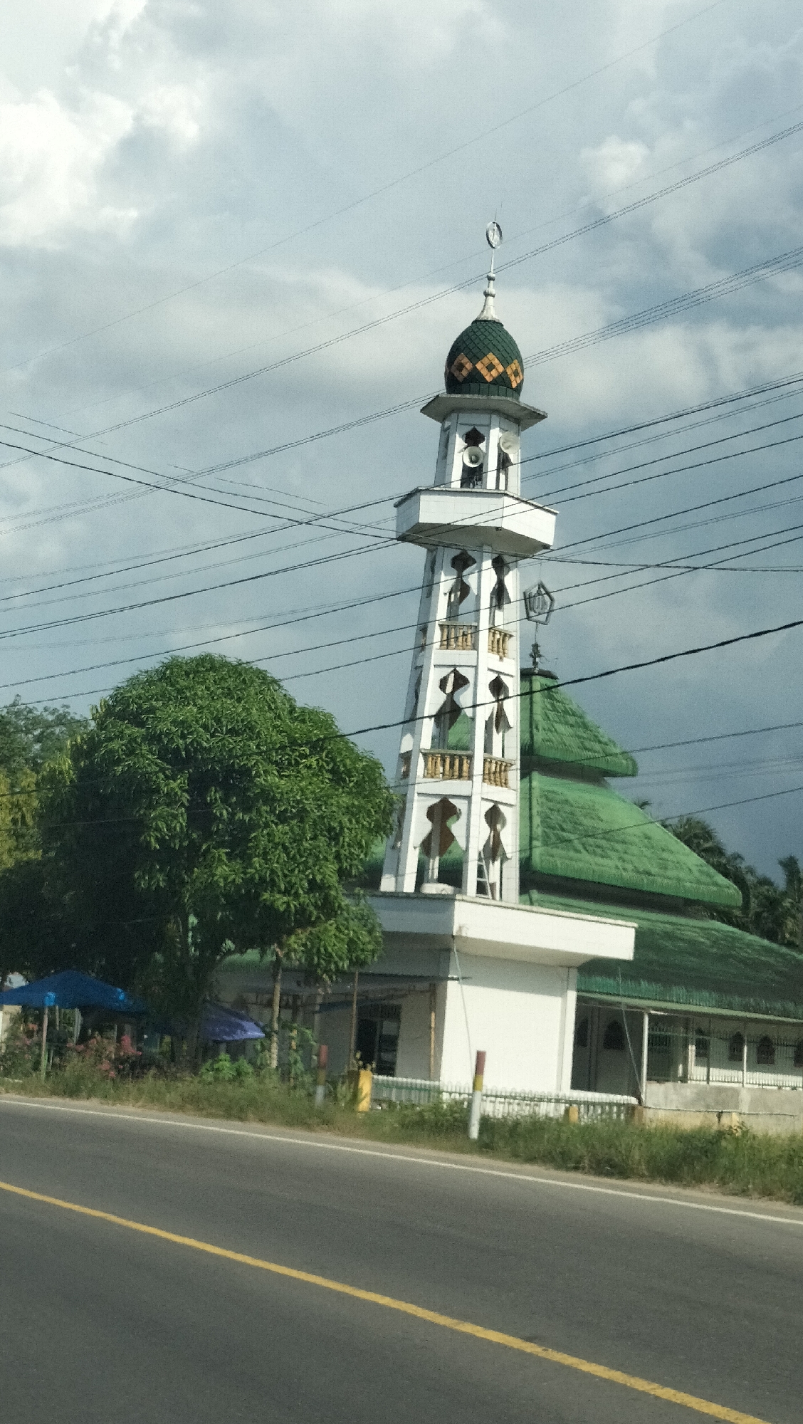 A mosque with a white tower and green roof is visible on the side of a road in Tangkahan Durian, Indonesia. The tower has several levels with arched openings and a crescent moon symbol at the top. A large green tree stands to the left of the mosque, and power lines run across the sky. The road in front of the mosque is paved and has a yellow line down the center. The sky is overcast with clouds.