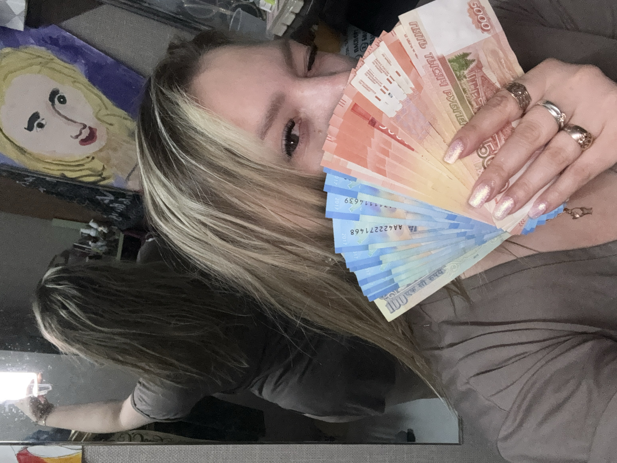 A woman is holding a fan of Russian rubles in front of her face. The bills are of different denominations, including 5000 ruble and 100 ruble notes. She has blonde hair with highlights and is wearing several rings on her fingers. In the background, there is a painting of a woman and other items on a shelf. The image appears to be a selfie taken in a mirror. The location in the city of Марковский, Russia cannot be determined from the image.