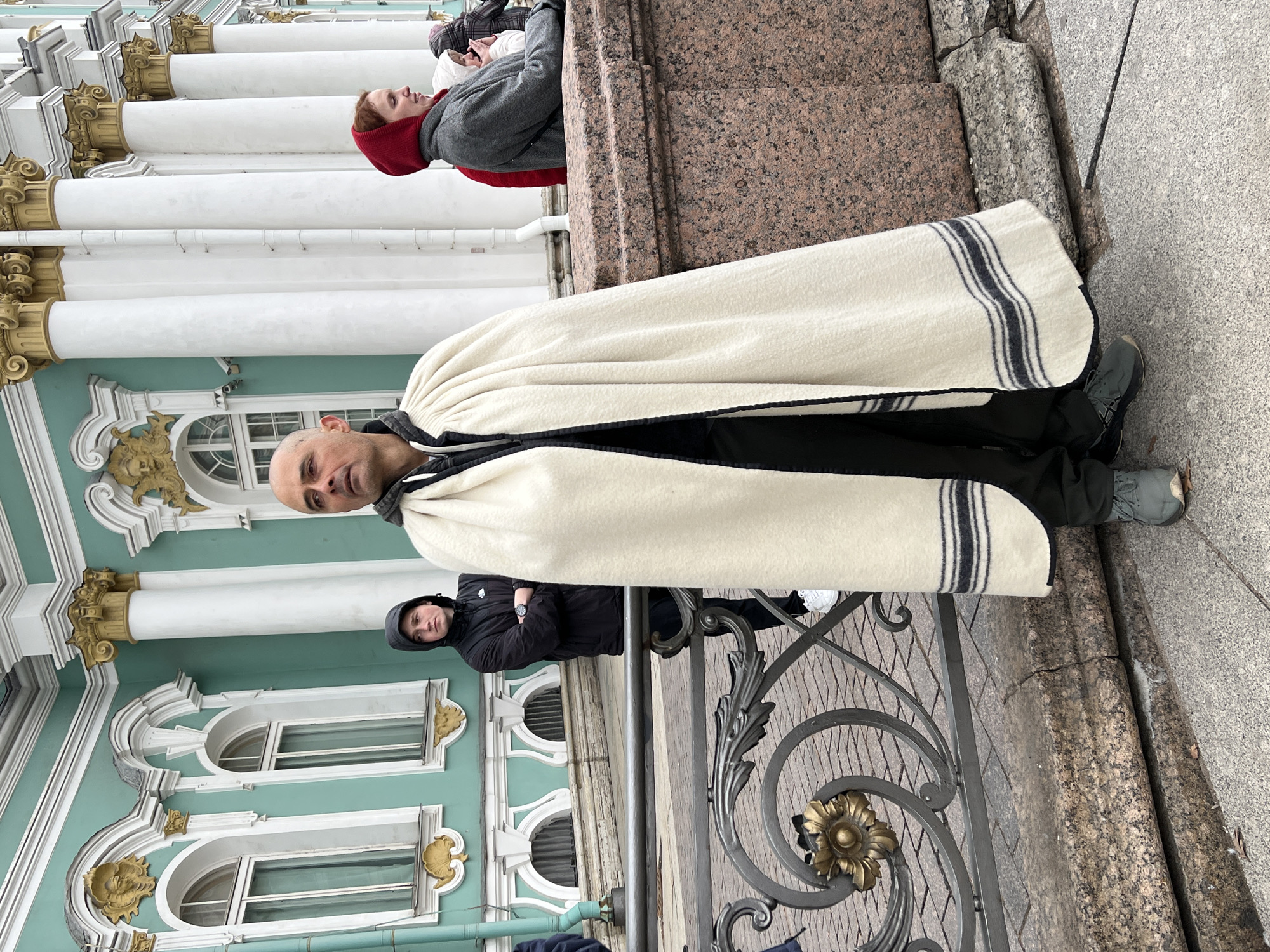 The image displays an outdoor scene in Saint Petersburg, Russia, featuring a grand, historic building as the primary backdrop. The building exhibits a distinctive mint green facade, adorned with tall, white fluted columns topped with ornate gold Corinthian capitals and decorative arched windows with gold accents, strongly indicative of the Winter Palace (Hermitage Museum).
In the foreground, a bald man with a neutral expression is captured looking directly towards the camera. He is clad in dark clothing, possibly black pants and a top, and is enveloped in a thick, cream-colored, blanket-like wrap featuring prominent black stripes and trim. He wears dark athletic shoes.
Behind him, slightly to the left, another individual with reddish hair, dressed in a grey hooded sweatshirt and a red inner hood, is seen leaning against one of the white columns, looking away from the camera. Further to the right and in the mid-ground, a third person wearing a black hooded jacket and white athletic shoes stands behind an ornate black metal railing decorated with gold floral patterns.
The ground consists of paving made from a mix of grey and reddish-brown granite stones, and a large reddish-brown granite block or pedestal is visible. The soft, diffused lighting suggests an overcast or cloudy day, implying potentially cool weather given the attire of the individuals. No discernible text is visible within the frame.