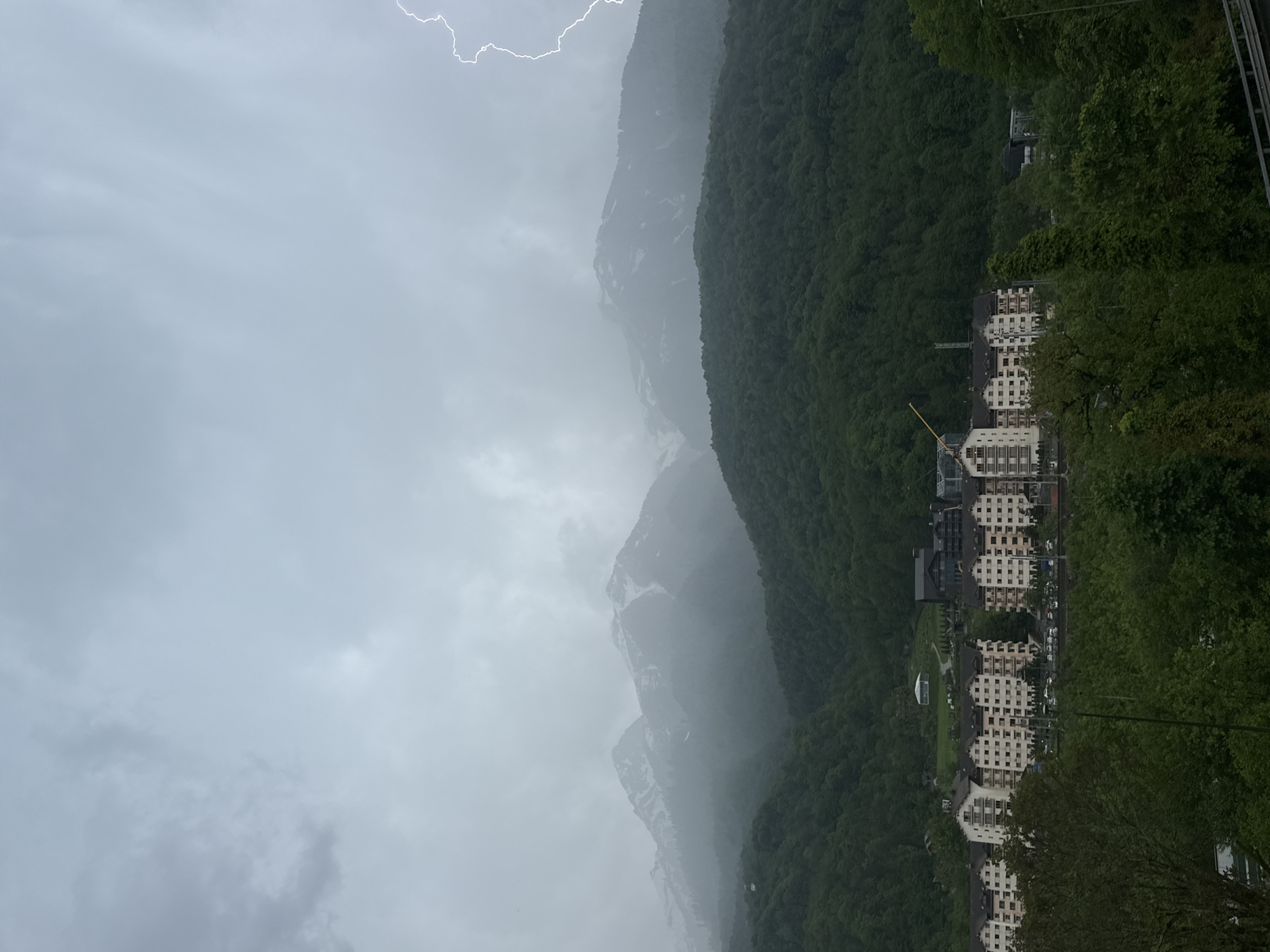 The image shows a mountain landscape in Krasnaya Polyana, Russia, under a cloudy sky with visible lightning. Mountains are in the distance, partially obscured by mist or clouds. Dense green forests cover the lower slopes. Buildings, likely hotels or residential complexes, are nestled among the trees, suggesting a resort or residential area in the valley. A construction crane is visible near one of the buildings.