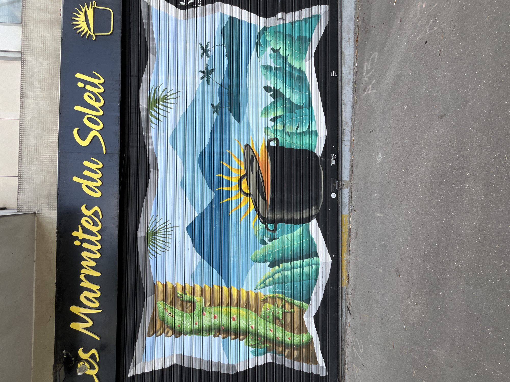 This media file shows a mural painted on a metal roll-up door, likely the storefront of a business. The mural depicts a tropical scene with vibrant colors. In the foreground, a bright yellow sun with a pot or cauldron in front of it is a central element. The pot appears to be boiling or emitting steam, hinted at by orange wisps. Surrounding the pot are lush green tropical leaves, some large and fan-shaped.
Below this, a yellow landscape suggests dry earth or sand. On this landscape, several green lizards with reddish spots are scattered. One lizard in the center is more prominently featured. In the background, a bright blue sky with stylized diamond shapes suggests mountains or a geometric interpretation of the sky. Palm trees with thin trunks and fronds are also visible, reinforcing the tropical theme.
To the left of the mural, a black sign with yellow, stylized text reads "Les Marmites du Soleil." Above this text is a yellow emblem that also appears to be a sun and a pot. The overall impression is cheerful and evokes warmth and perhaps a connection to cooking or food, given the name of the establishment. The scene is brightly lit, suggesting daytime. The setting appears to be an urban environment, as indicated by the asphalt road visible to the right of the door.