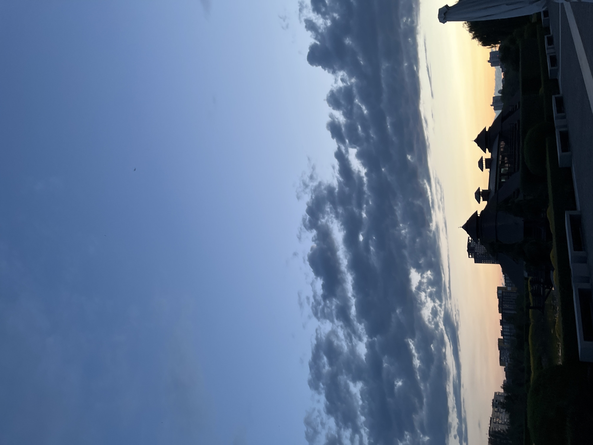 The image captures a twilight scene in Dnipro, Ukraine. The sky is a gradient of soft blues and yellows, transitioning from a clear, pale blue on the left to a warm, sunset-tinged horizon on the right. A line of dark, silhouetted buildings marks the cityscape along the horizon, with a few trees interspersed. In the foreground, silhouetted structures, possibly part of a modern building complex with distinctive rooflines and turrets, are visible against the vibrant sky. The clouds are dramatic, with dark, textured formations stretching across the upper portion of the sky, contrasting with the calmer, clearer blue. The overall impression is one of a serene evening in an urban setting, with the light of dusk casting long shadows and highlighting the architectural features of the buildings. There are no people visible in the image. The time of day is clearly twilight, just before nightfall.