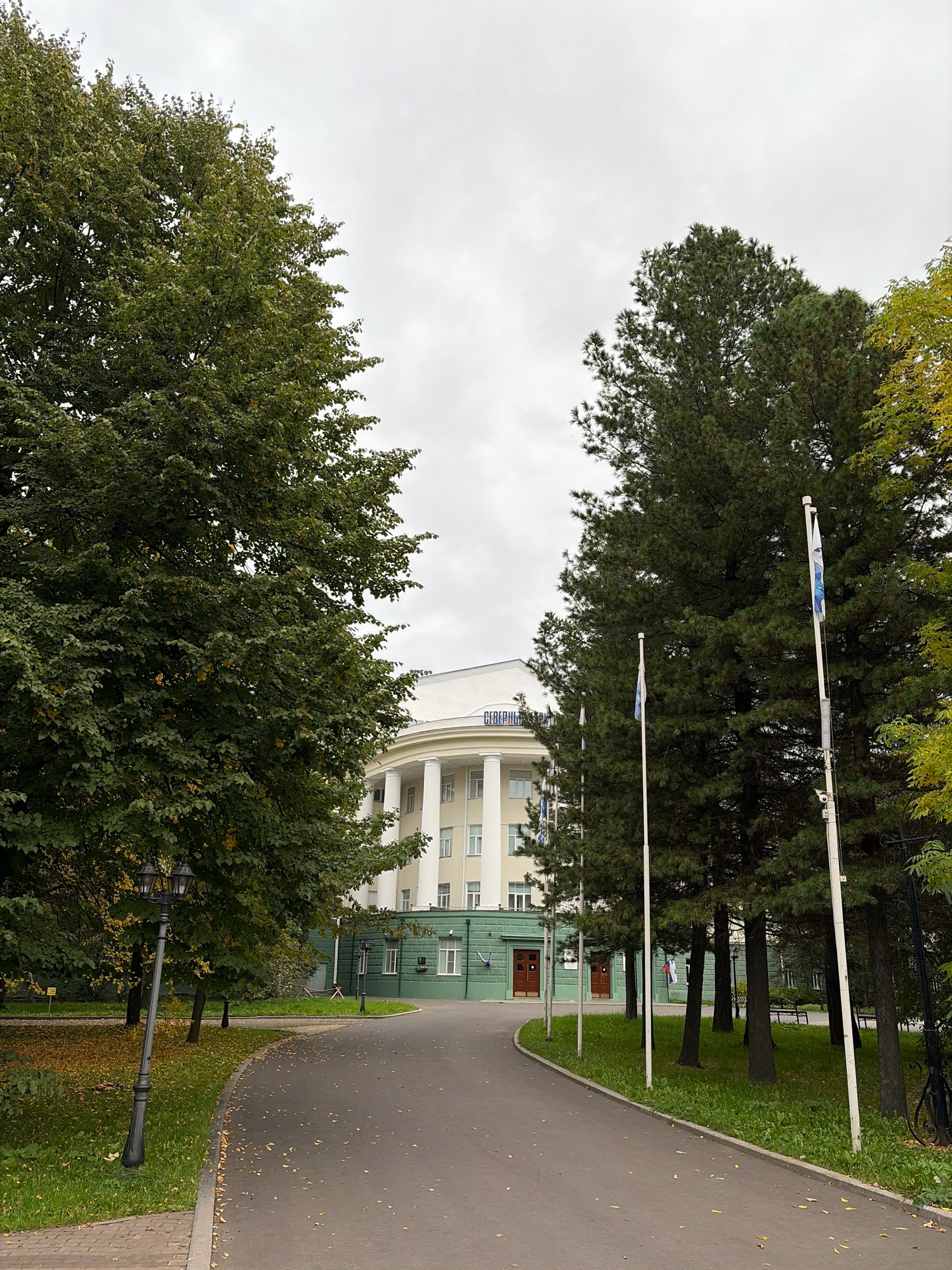 The image depicts the Arkhangelsk State Technical University, a notable building in Arkhangelsk, Russia. The foreground features a paved walkway curving to the right, flanked by a grassy area with fallen leaves and a vintage-style lamppost on the left. Tall evergreen trees stand on the right side of the walkway, interspersed with flagpoles flying flags. In the background, a grand, light-colored building with a semi-circular facade is visible, characterized by large white columns and multiple windows. A green section with doors is situated at the base of the building. The sky is overcast, suggesting a cloudy day.