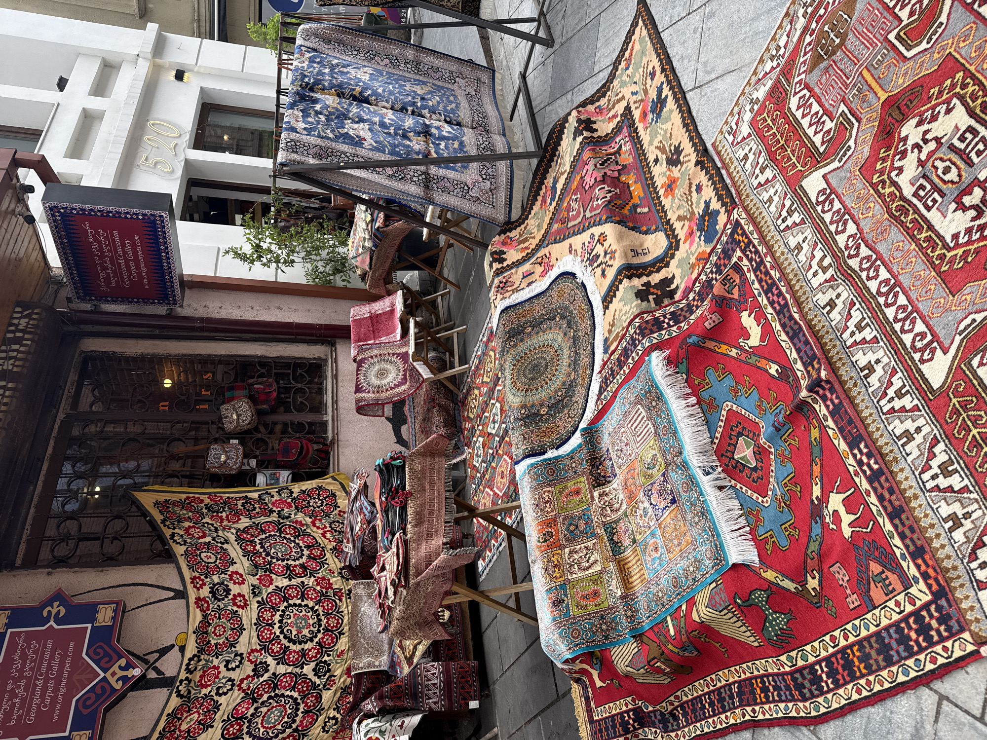 The image shows a street scene in Tbilisi, Georgia, featuring a carpet gallery. Various carpets are displayed on stands and the ground, showcasing intricate patterns and colors. Signs advertise the gallery as "Georgian & Caucasian Carpets Gallery" with the website "www.origincarpets.com" indicated. The number "520" is displayed on a building in the background, and a variety of handbags are visible in the storefront. The environment suggests a commercial district or marketplace.