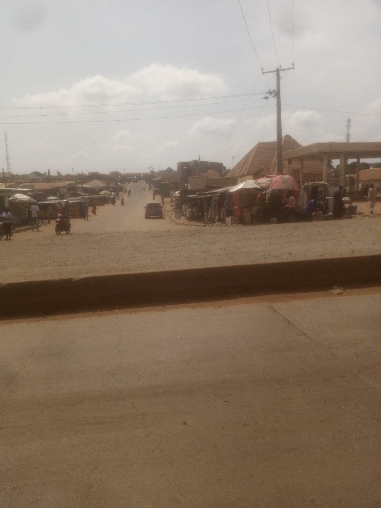 The image depicts a bustling street scene in Nassarawa, Nigeria, under a hazy sky with scattered clouds. The foreground shows a dirt or gravel road or pathway, with a concrete barrier in front of it. The middle ground features a street lined with various structures, including market stalls with awnings and shelters, some appearing to be vendors selling goods. There are several parked vehicles, including what appear to be auto-rickshaws and motorcycles. People are seen walking, standing, and engaged in activities associated with a marketplace. In the background, more buildings are visible, along with utility poles and power lines. A gas station with a distinctive roof structure is also present on the right side of the image. The overall impression is of a busy, typical urban environment in Nigeria.