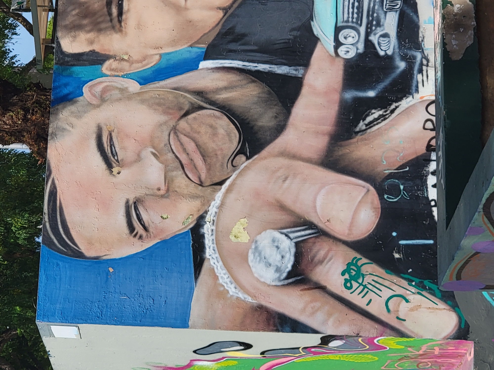 This is a close-up photograph of a mural painted on a concrete wall in Quarteira, Portugal. The mural depicts two stylized portraits of men, rendered in a realistic but somewhat exaggerated style. The primary focus is on the left-hand figure, who is shown with his head turned and his tongue sticking out, making eye contact with the viewer. He is wearing a large, ornate ring with a prominent, light-colored stone on his right index finger. The ring has a textured band. A small, green graffiti tag, resembling a stylized spider or insect, is visible on the index finger below the ring. The man on the right is partially visible, with his head and shoulder also painted. The background of the mural is a vibrant blue, contrasting with the skin tones of the men. A portion of a light blue vintage car, with visible headlights and grille, is depicted in the lower right corner. The mural shows signs of wear, with peeling paint and chipped sections on the concrete surface, particularly around the edges and on the blue background. The overall scene suggests an outdoor urban environment, possibly a public space or skate park, given the presence of graffiti and the style of the artwork.