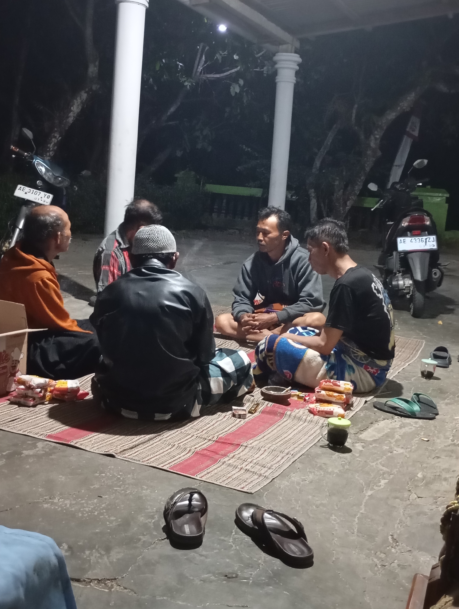 This photograph captures a nighttime gathering of five adult males seated on a mat on a concrete floor, likely in an outdoor or semi-outdoor setting in Wonogondo, Indonesia, adjacent to a building with prominent white pillars. The lighting suggests artificial illumination, creating shadows.
From left to right, a man in an orange shirt and black pants is seated with his back to the camera, facing inward towards the group. Next to him, another man, partially visible, wears a red and grey plaid shirt. The central figure, facing away from the viewer, is dressed in a black leather jacket and a white patterned cap, with a patterned sarong wrapped around his waist. To his right, a man in a grey hoodie and patterned shorts is seated with his hands clasped. The fifth man, on the far right, wears a black t-shirt with a graphic design and patterned shorts.
Scattered around the mat are several objects. A cardboard box, partially visible on the left, contains packets of what appear to be snacks. A small dish or ashtray sits near the center of the mat, along with several cigarette packs and a lighter. A glass with a green lid, containing a dark liquid, is also present. Two pairs of flip-flops are visible on the concrete floor, one dark brown and the other dark green.
Two motorcycles are parked in the background. The motorcycle on the left has a license plate reading "AE 3107 YD". The motorcycle on the right has a license plate reading "AE 4336 TJ". Trees and foliage are visible behind the motorcycles and the pillars, suggesting a somewhat natural environment surrounding the gathering space. The overall impression is a casual, communal gathering taking place after dark.