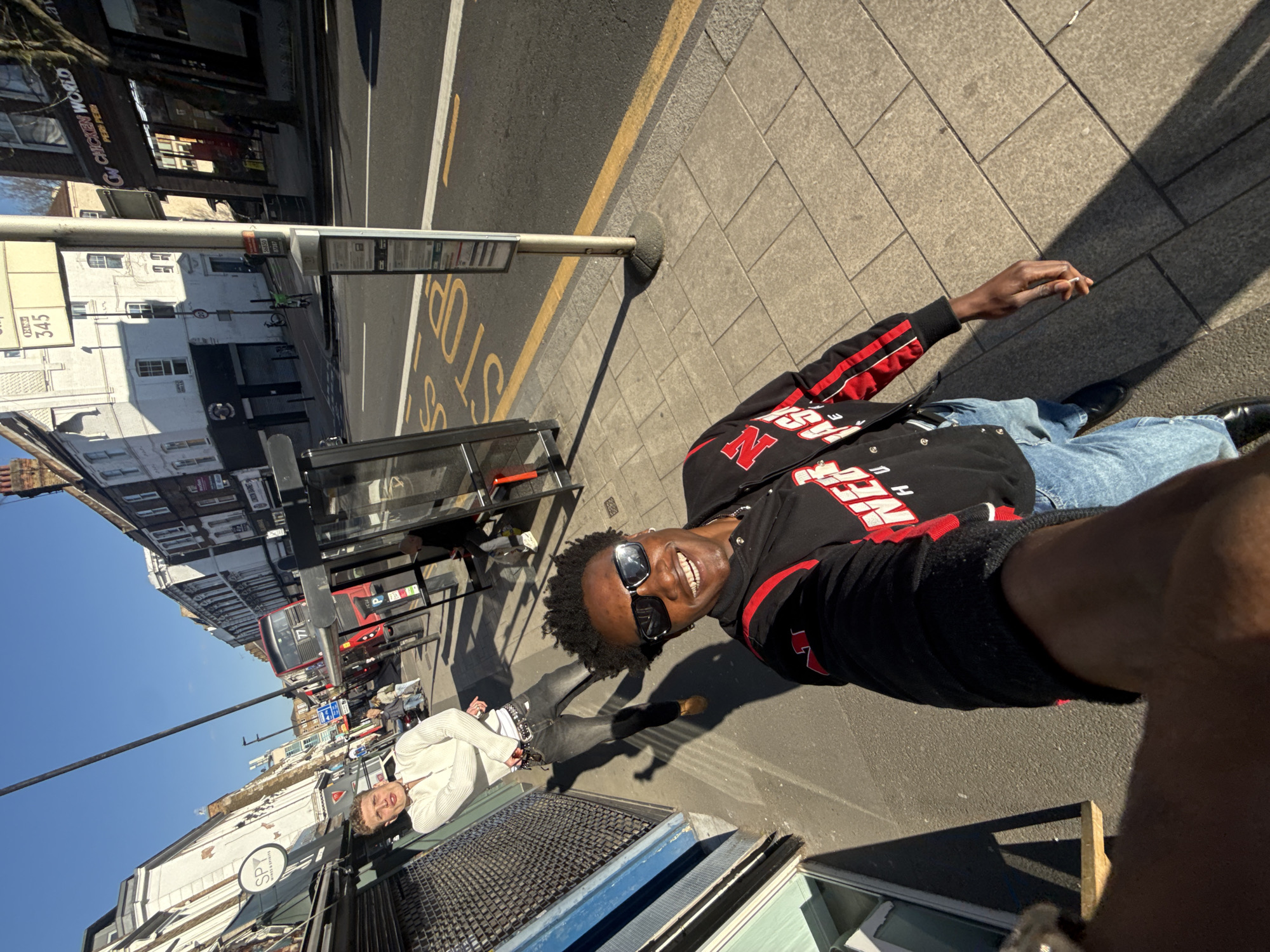The image shows an individual taking a selfie on a sunny day in London, United Kingdom. The person is wearing a black jacket with red and white lettering, blue jeans, and sunglasses, and is smiling at the camera. They are standing on a sidewalk next to a bus stop, with the word "STOP" painted on the road. In the background, there are buildings, shops, and a red double-decker bus. A second person wearing a white sweater is standing in the background as well. One building in the background has a sign that reads “Chicken World”. The image is rotated.