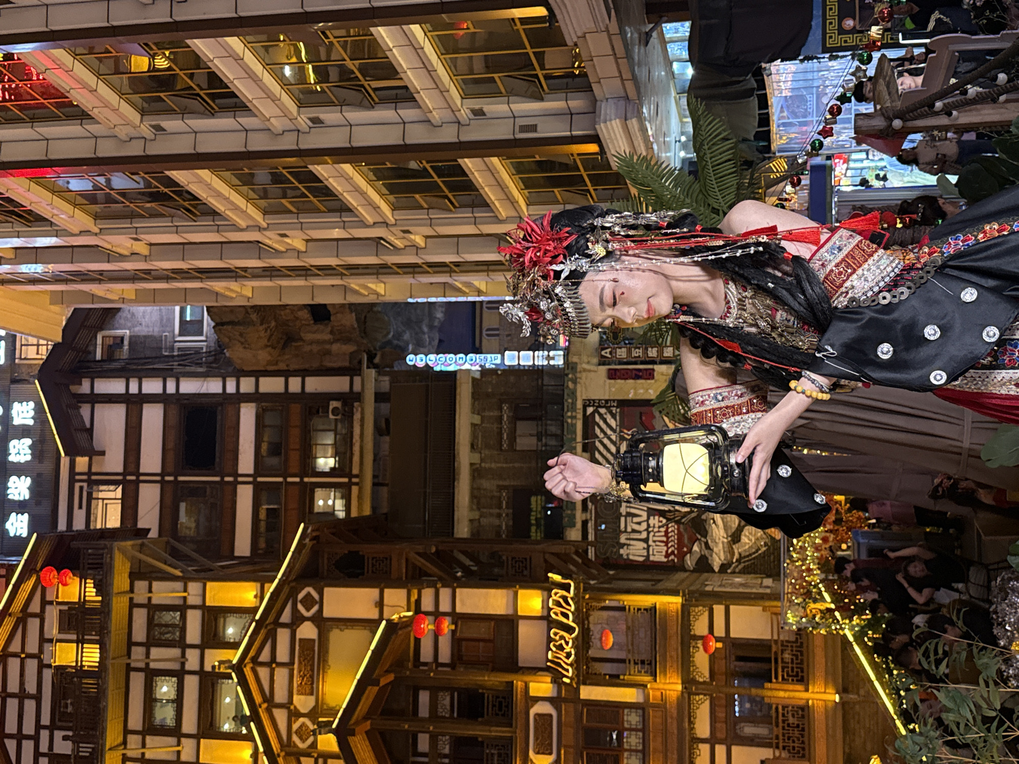 The image depicts a vibrant night scene in what appears to be a historic or culturally themed district, likely in Yuzhong District, China. The primary subject is a woman centrally positioned, elegantly dressed in an elaborate traditional costume. Her attire features intricate embroidery in red and gold patterns, black fabric embellished with silver button-like elements, and long flowing sleeves. She wears an ornate headpiece adorned with red flowers, silver ornaments, and dangling beads. Her face, seen in profile looking downwards, shows serene or pensive expression, with a small red dot on her forehead. She holds a vintage-style lantern with a warm glow in her right hand.
The setting is dominated by multi-story traditional Chinese-style buildings with distinctive wooden frameworks, white walls, and layered roofs. These structures are beautifully illuminated by warm yellow string lights that outline their architectural features, creating a festive atmosphere. Several red lanterns hang from the eaves of the buildings. In the background, other buildings are visible, some with lit windows. Faint glimpses of other people can be seen in the lower right, suggesting a busy public area. Green foliage from potted plants or landscaping is visible on the sides. The time of day is clearly night, evidenced by the darkness and reliance on artificial lighting.
Visible text includes Chinese characters on signs. On a building in the upper left (when the image is properly oriented), vertical characters appear to include "银楼" (Yínlóu - Silver Building/Store). On a sign near the bottom left, stylized horizontal characters are visible, possibly "火锅" (huǒguō - hotpot) or a similar establishment. Other faint Chinese characters are present on various signs in the background.