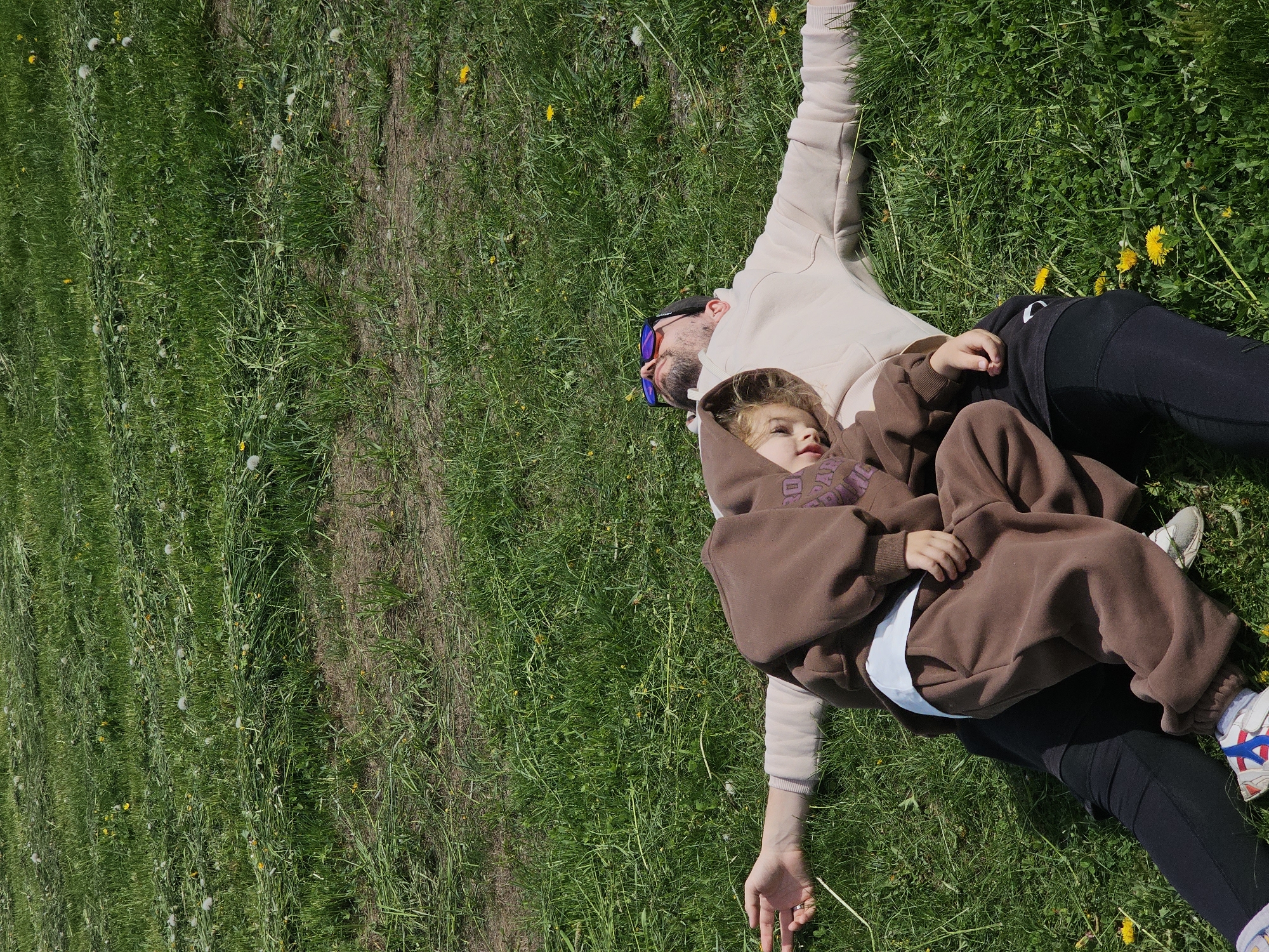 A top-down shot shows a man and a child lying on a patch of green grass dotted with yellow dandelions and white dandelion seed heads. The man is wearing a beige hoodie, black leggings, and blue-tinted sunglasses, with his arms outstretched. The child is nestled on top of the man, wearing a brown hoodie over a white shirt, black leggings, and sneakers with red and blue accents. The child's hands are resting on their stomach. The grass appears to be recently mowed with uncut patches. The image appears to be taken in daylight. The specific location within Планета Снт, Russia is not discernible from the provided image.