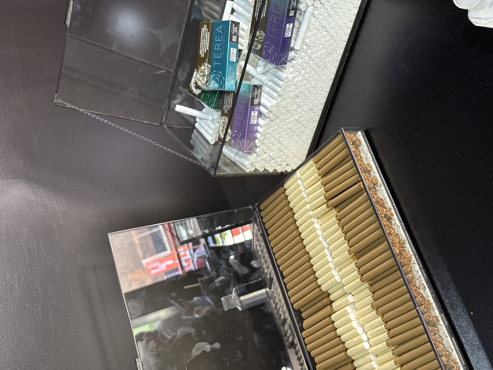 The image shows two acrylic containers filled with cigarettes and other related products, likely taken at a retail location. One container holds loose white cigarettes and packs of "TEREA" brand tobacco sticks for IQOS devices. The other container holds brown and white Chapman brand cigarettes, arranged in rows. The reflection in one container shows a storefront, possibly in Yerevan, Armenia, although this can only be inferred from the appearance of the store's signage. No people are visible in the image. The scene captures a commercial display of cigarettes and tobacco products.
