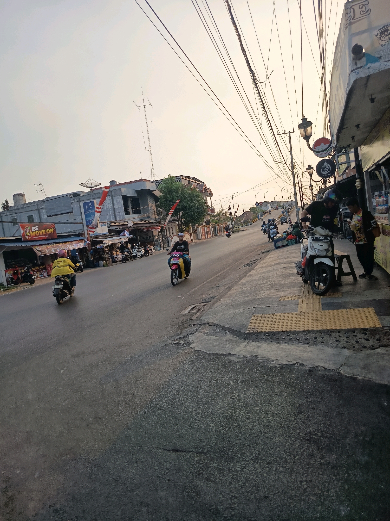 The image depicts a street scene in Banjarmasin, Indonesia, under an overcast sky. On the right side of the frame, a row of small businesses lines the sidewalk, featuring various signage and merchandise displayed. One business appears to be a food stall with bottled drinks visible. A white motorcycle is parked on the sidewalk next to a bench, with a person on it wearing a black t-shirt and helmet. Another person, wearing a colorful patterned shirt and black pants, stands beside the motorcycle.
The street itself is paved and appears wet, indicating recent rain or high humidity. Several motorcycles are in motion on the road. In the foreground, a person in a yellow jacket and helmet rides a dark-colored scooter. Further down the street, another person in a pink and yellow outfit is also riding a scooter. In the background, more motorcycles and possibly a car are visible.
The buildings on the left side of the street are a mix of commercial structures, with one featuring a prominent sign that reads "ES MOVE ON". Indonesian flags are displayed on poles attached to some of the buildings. Utility poles with numerous wires are a prominent feature of the urban landscape. A satellite dish is mounted on the roof of one of the buildings. The overall impression is of a busy, functional urban environment.