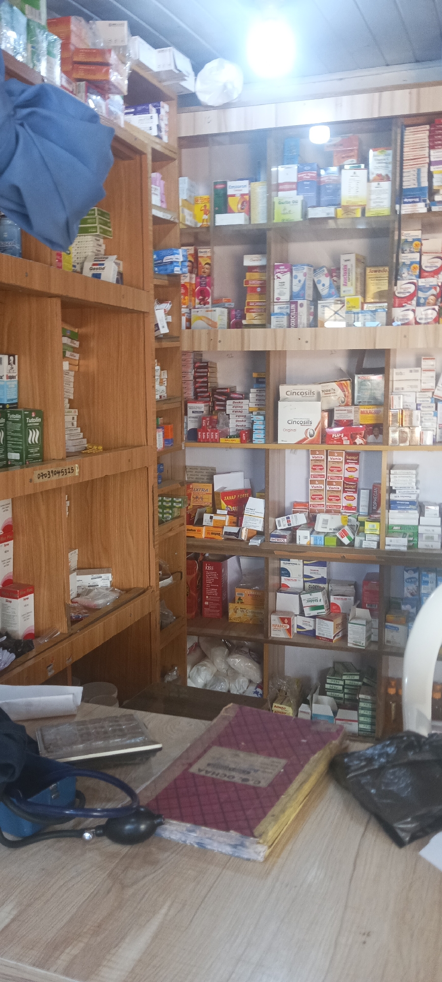 This is an interior shot of what appears to be a pharmacy or medical shop, likely in Kuru Dungus, Nigeria. The shelves are made of light-colored wood and are packed with various medications, pills, and other medical supplies. Brands such as "Cincosils," "ROPHEGAN," "MOXICILLIN," "Jowado," "XANAP FORTE," "Gestid," and "RIFABION" are visible. A number, likely a phone number, "07039045325" is also visible. In the foreground, there's a wooden counter with a notebook, a blood pressure cuff, and a black plastic bag. The lighting is bright.