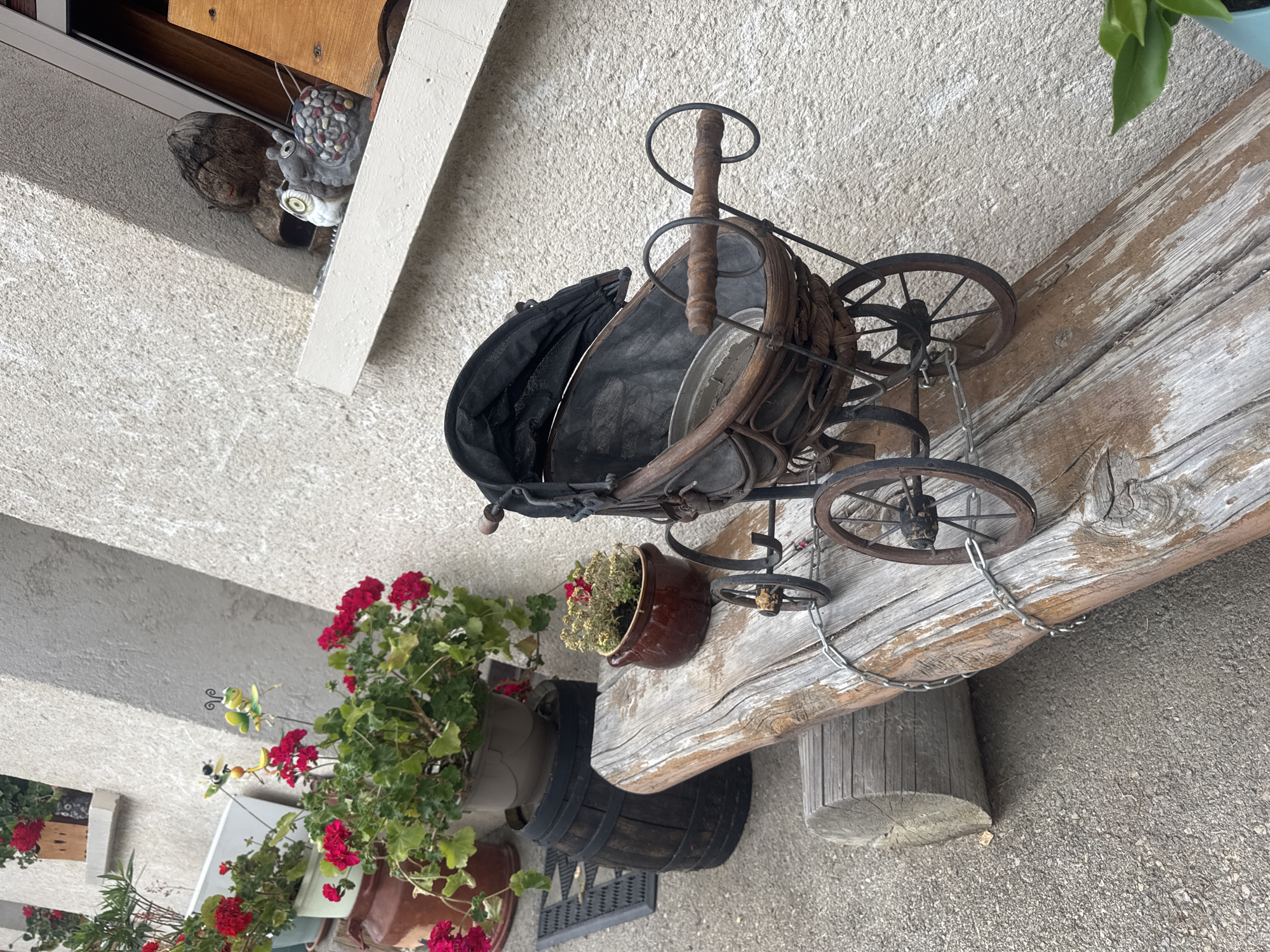 The image depicts a rustic outdoor scene in Les Rousses, France, likely a residential area or garden. The foreground features a weathered wooden beam serving as a bench, with a small, antique-style doll carriage placed upon it. The carriage is made of dark wood with a fabric canopy and metal wheels, secured by a chain. To the left of the bench, a dark wooden barrel is visible, topped with a pot containing red flowers and greenery. Further left, a doormat and more potted plants with red flowers are present. Behind the bench and barrel, a textured, light-colored building wall forms the background. On a windowsill above and to the right of the carriage, several decorative figures are displayed, including two owl figurines and a snail. The overall impression is one of a charming, slightly aged, and decorative outdoor space.