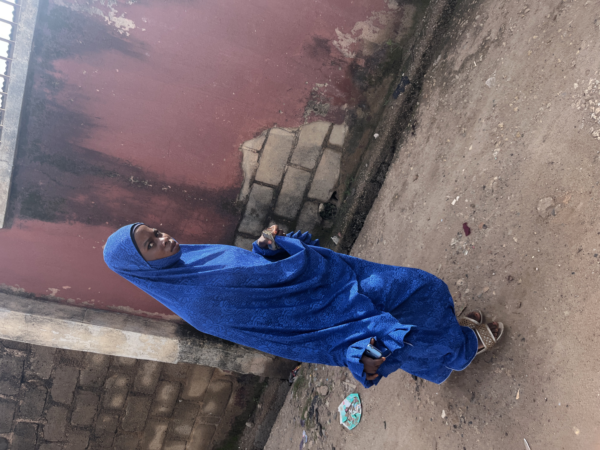 A young woman is photographed from a slightly elevated perspective, standing in what appears to be an outdoor urban environment. She is wearing a vibrant royal blue headscarf and a matching floor-length dress with a subtle, repeating floral pattern. Her attire suggests modest dress, common in many Nigerian cultures. The woman is looking upwards and slightly to her left, with a neutral expression on her face. Her right hand is raised, holding a small item, possibly a phone or a wallet. Her left hand is visible near the hem of her dress, holding what also appears to be a phone. She is wearing open-toed sandals with a woven strap design.
The setting behind her is characterized by a textured, reddish-pink wall that shows signs of weathering and some patches of dirt and grime. Below the wall, there are glimpses of brickwork and a rough, dusty ground surface scattered with small stones and debris. To the left of the frame, a section of a grey, possibly concrete, structure with metal bars is visible, suggesting a building or boundary. The ground is a mixture of packed dirt and gravel. The lighting indicates it is daytime, likely with clear skies, casting subtle shadows. The overall scene evokes a sense of everyday life in a possibly developing urban area. There is no visible text in the image.
