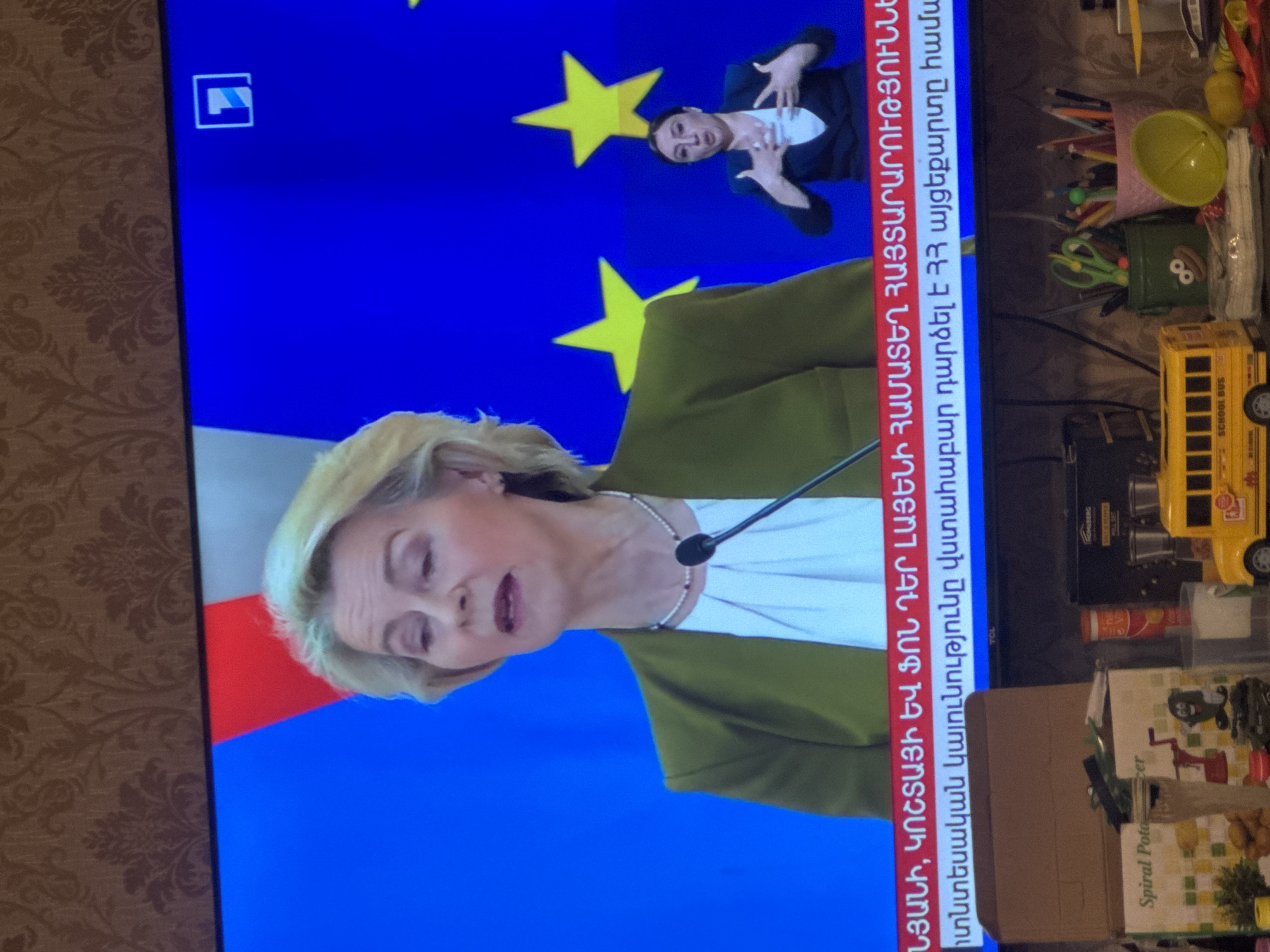 The image shows a television screen displaying a news program. The program features Ursula von der Leyen, President of the European Commission, and an interpreter for the deaf who is signing in the top right corner. The background is a flag of the European Union with yellow stars on a blue field. Text in Armenian is on the right side of the screen. In front of the television, there is an assortment of objects including stationery, a school bus toy, and a box labeled "Spiral Potato." The location appears to be in a home setting, likely in Yerevan, Armenia, given the Armenian text on the screen.