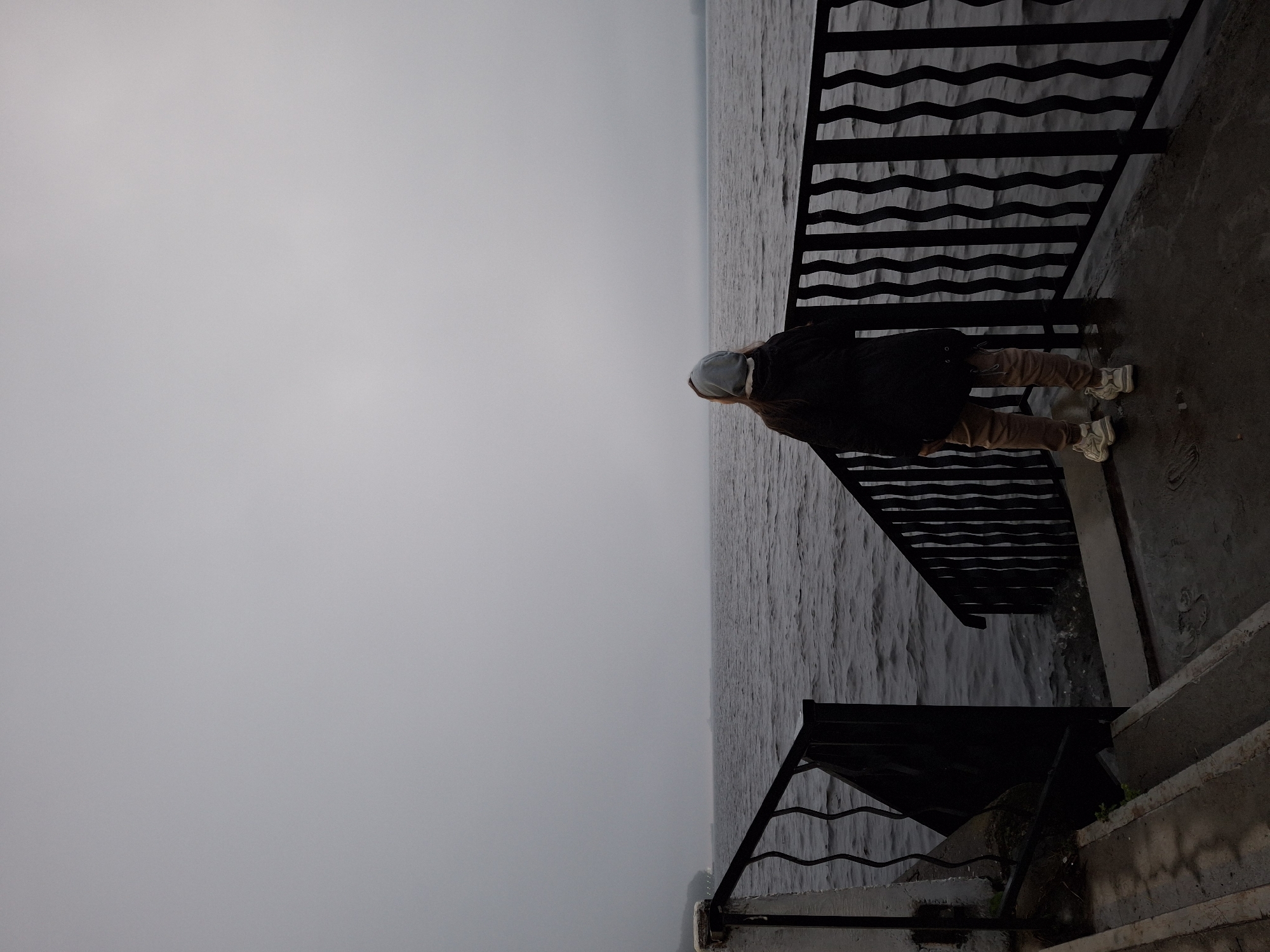 A person stands on a concrete platform, looking out at a body of water under an overcast sky. The person is wearing a light blue hood, a dark jacket, and tan pants, with white sneakers. A black metal railing with a wavy pattern runs along the edge of the platform. Stairs are visible to the left and right of the platform, leading down to the water. The location appears to be a waterfront promenade or pier, possibly in Chaikovsky, Russia, given the presence of a large body of water and a somewhat industrial or utilitarian waterfront design. The water is choppy, suggesting wind. The overall atmosphere is somber and quiet, consistent with a cloudy or misty day.