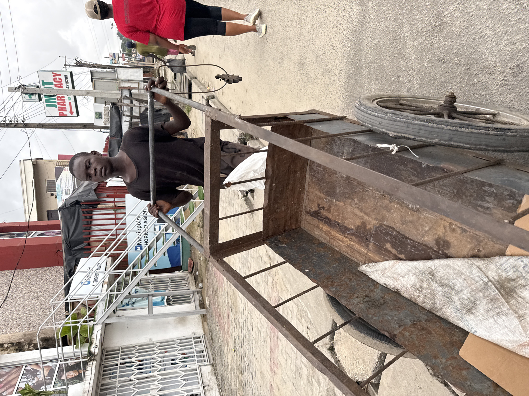 This street scene, likely in Ado, Nigeria, captures a young man standing with a large, rustic handcart under an overcast sky. The man, wearing a dark short-sleeved shirt, holds the metal handles of the sturdy, well-used cart, which features a rusty metal frame and a wooden base. A white sack or bag is visible inside the cart's bed. One wheel of the cart is prominent, showing wear and tear.
In the background, another person, dressed in a red shirt and black shorts, walks away from the viewer on what appears to be a paved sidewalk or road. Utility poles with numerous wires crisscross above. Several buildings line the street, some with visible shopfronts and signage. A white metal staircase or railing is visible on the left side of the frame, leading up to a building entrance.
Notable visible text includes a prominent green and white sign that reads "JMT PHARMACY" accompanied by a cross symbol, and a phone number "080 4218205". Another sign, partially obscured, appears to belong to "M-Mobilepoint" and "AUTHORISED AGENT". The overall setting suggests a bustling urban environment with commercial activity. The time of day appears to be daytime, with soft, diffused light indicating an overcast sky.