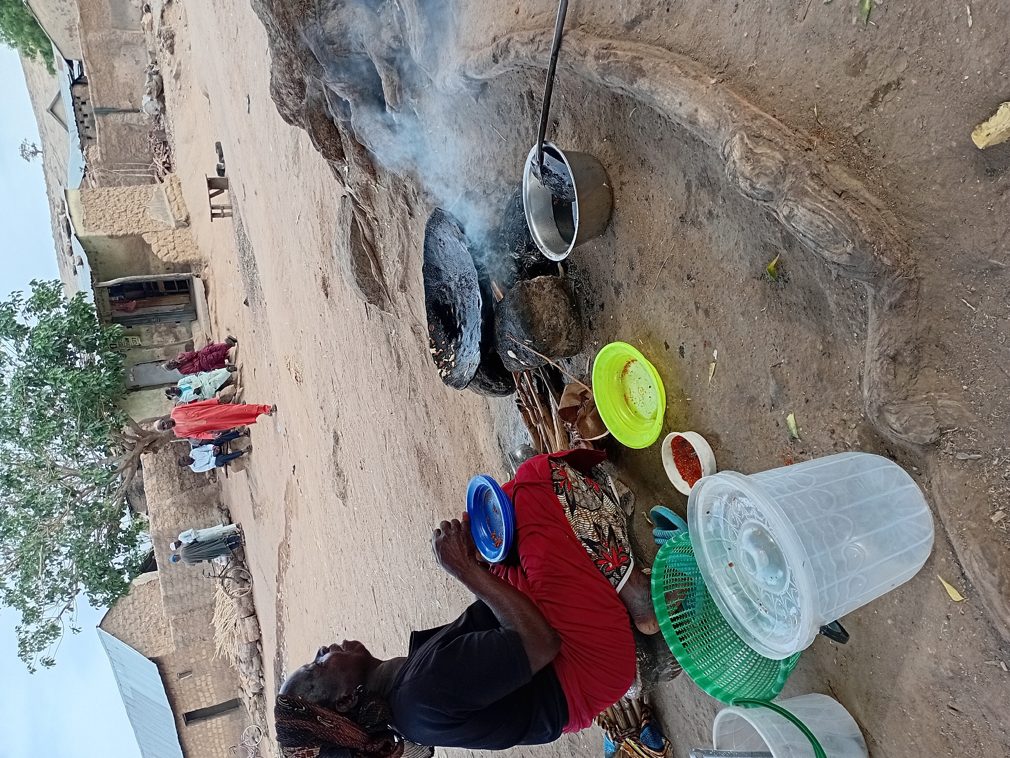 An image depicts a woman seated on the ground next to an outdoor cooking area in Nata-Ala, Nigeria. The cooking area features a fire pit with smoke rising from it. At least two pots rest on or near the fire. The woman is dressed in dark clothing and a red skirt, and she is holding a blue plate. Around her are various plastic containers, plates, and baskets. In the background, there are several buildings and a small group of people standing near a tree.