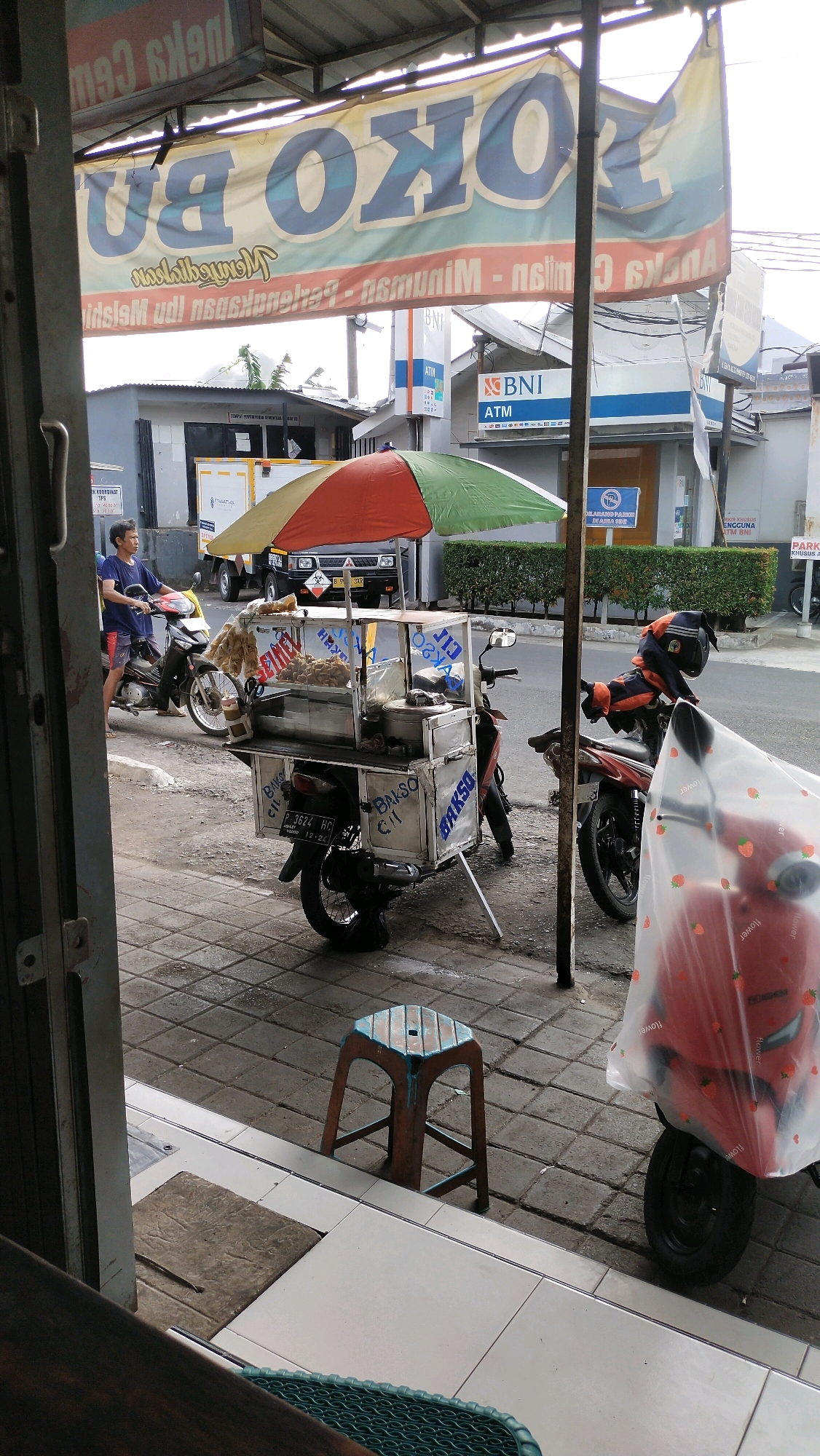 This image captures a street scene in Jember, Indonesia, likely during the daytime. In the foreground, a food cart modified from a motorcycle is parked on a tiled sidewalk. The cart has a red, yellow, and green umbrella providing shade over its display of food, which appears to be Indonesian meatballs ("bakso"). The motorcycle is decorated with text that reads "BAKSO CI." A man in a blue t-shirt and shorts is seated on another motorcycle to the left of the food cart, looking towards the cart. In the background, a street is visible with several buildings, including a Bank BNI ATM. The architecture suggests a commercial or urban area. A red motorcycle covered with a plastic sheet, adorned with strawberry prints, is parked on the right side of the frame. A small, rustic stool with a worn surface is placed on the sidewalk between the food cart and the covered motorcycle. The overall impression is a snapshot of daily commerce and street life in Jember.