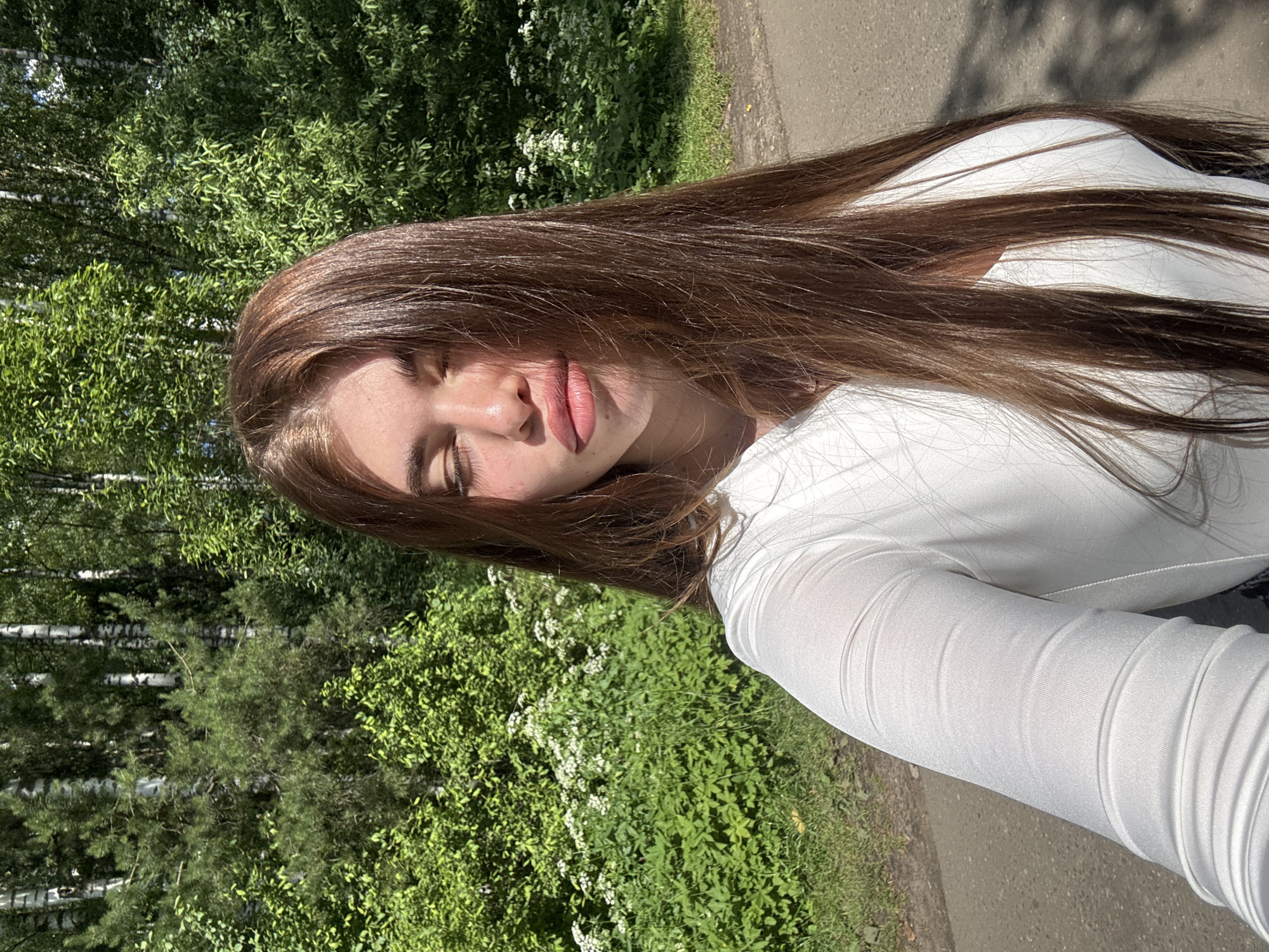 A young woman with long, brown hair is taking a selfie outdoors. She is wearing a white long-sleeved shirt. Her eyes are closed, and her lips are slightly parted. She is standing on a paved path, with a lush green forest and a patch of white wildflowers behind her. The sun is casting shadows on the path, indicating it is daytime. The scene appears to be in a park or natural area.