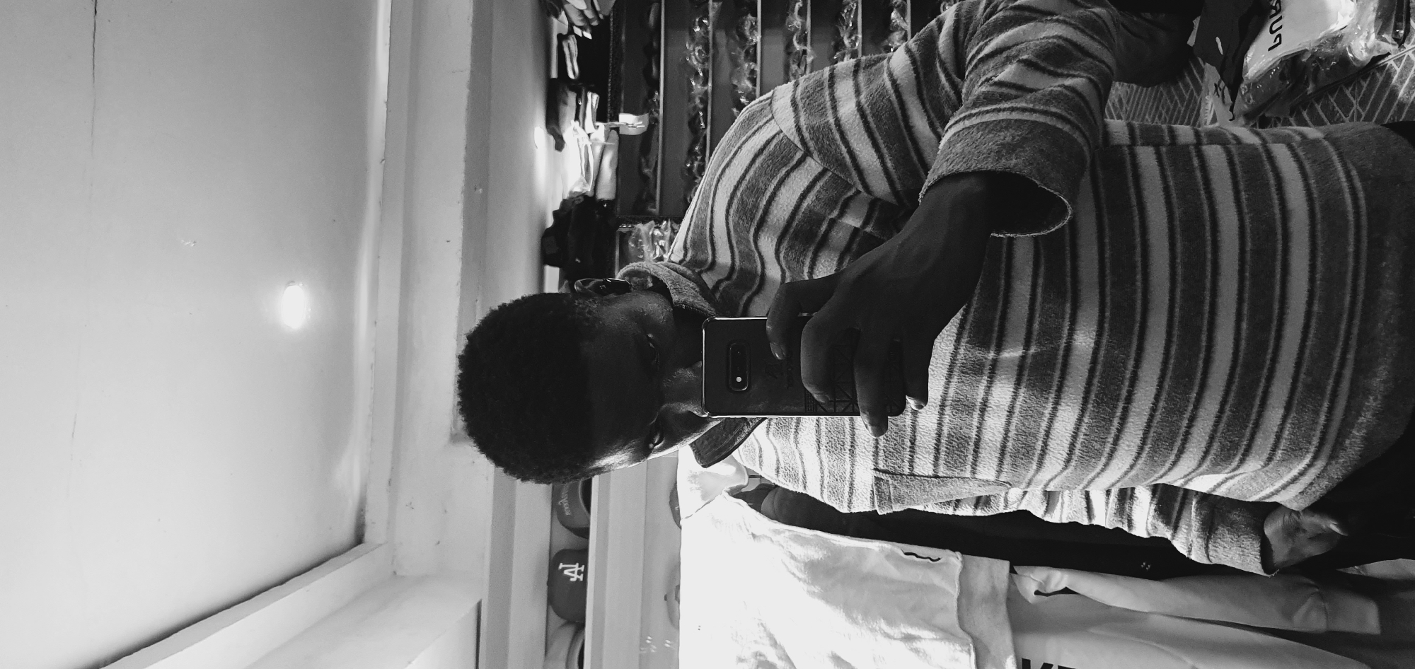 A black and white, waist-up, self-portrait photograph is taken by a young man in Bukuru, Nigeria. He is holding a smartphone in his right hand, angled to capture his reflection. The man is wearing a striped, collared sweater. Behind him, shelves are visible, filled with various items, including what appear to be hats and folded clothing. The room has a white ceiling with an illuminated light fixture. The overall impression is of an informal, personal moment captured in what seems to be a retail or storage space.