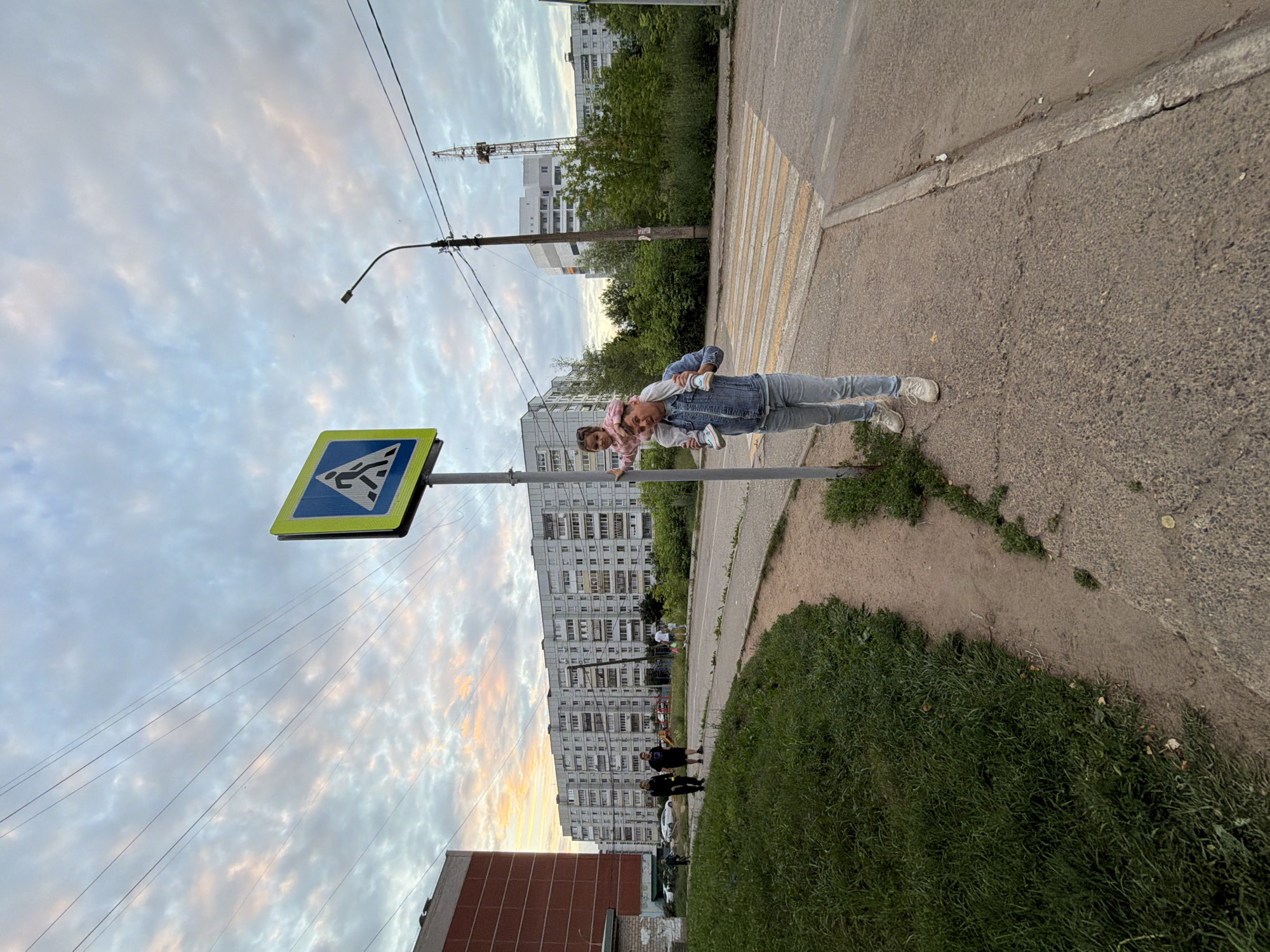 The image shows a man carrying a young child on his shoulders, standing on a sidewalk next to a pedestrian crossing sign in Chaikovsky, Russia. The man is wearing a denim jacket and light-colored jeans, and the child is wearing a pink top and pants, with a pink hat. In the background, there are multi-story apartment buildings typical of Soviet-era architecture, suggesting a residential area. The sky is cloudy with hints of sunset colors. Other people are visible walking in the distance near the apartment buildings. A crane is also visible in the far background, indicating ongoing construction or industrial activity. The overall scene depicts a moment in a typical urban environment during twilight.