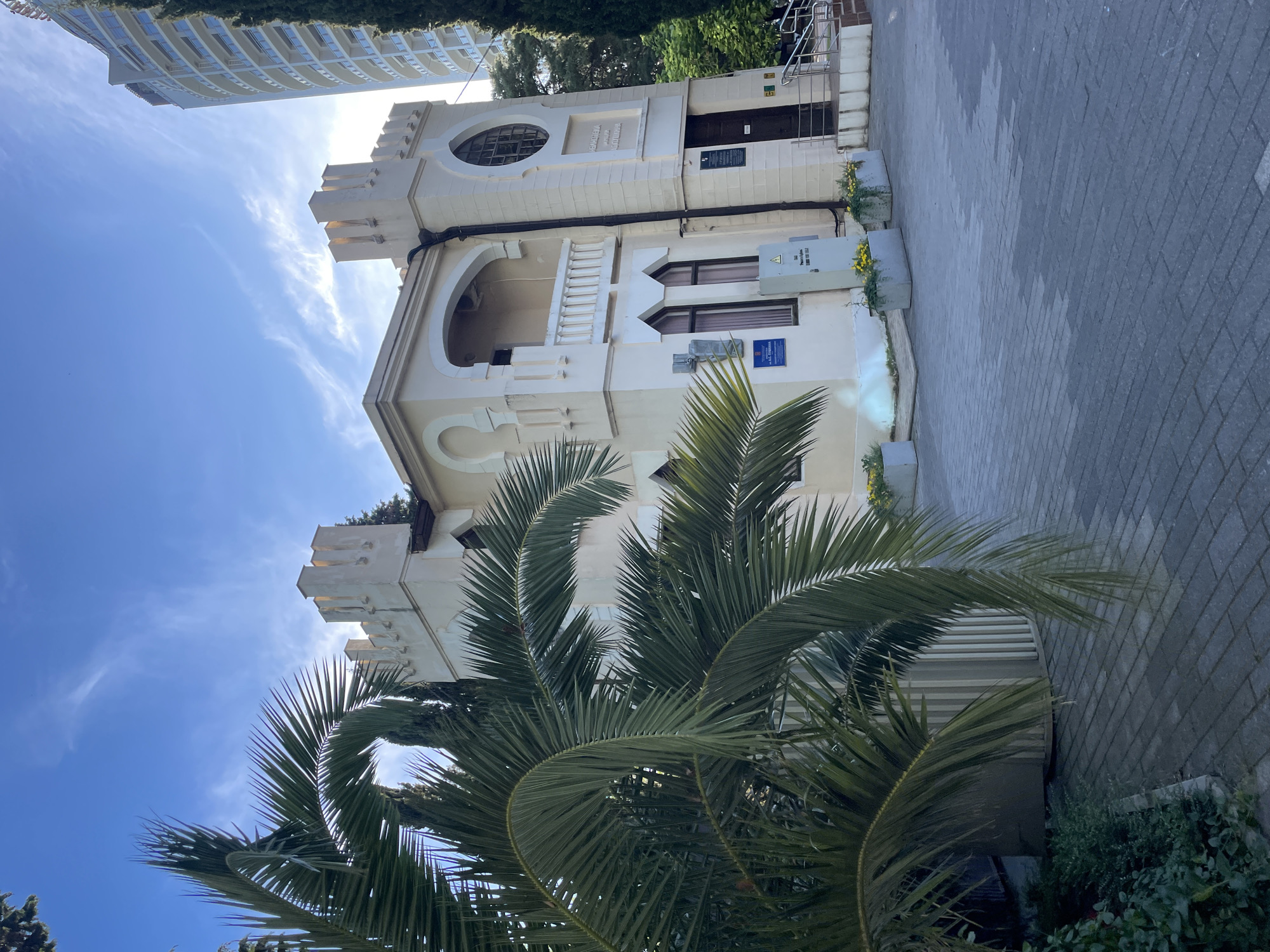 The image depicts a light-colored, two-story building with a crenellated turret, reminiscent of a small castle, situated in Sochi, Russia. The architecture suggests a historical building, possibly a museum or library, given the signs visible on its facade. In front of the building, a paved courtyard is visible, with large planters containing yellow flowers and greenery. A large palm tree with prominent fronds dominates the left side of the frame, partially obscuring the building. To the right of the building, a modern, tall, high-rise apartment building with balconies is visible, contrasting with the older architecture. The sky is blue with scattered wispy clouds, and the lighting suggests it is late afternoon. The scene appears to be an exterior view of a public space.