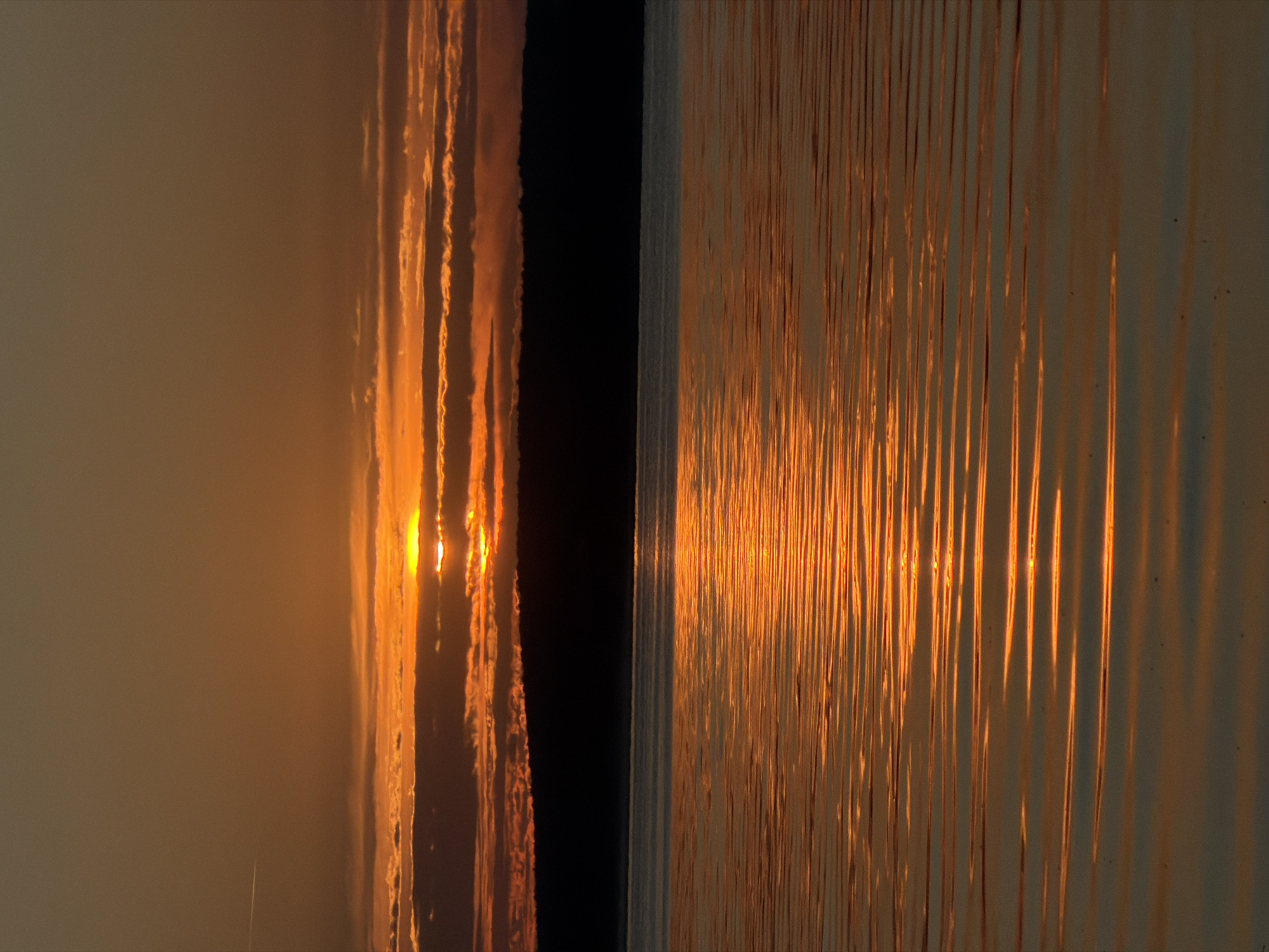The image depicts a sunset over a body of water, likely the Kama River, in Chaikovsky, Russia. The sun is partially obscured by clouds, casting an orange and gold glow across the sky and reflecting on the rippling water. A dark, silhouetted treeline is visible in the distance, separating the sky from the water. There are no people or distinct objects in the scene, which focuses solely on the natural landscape.