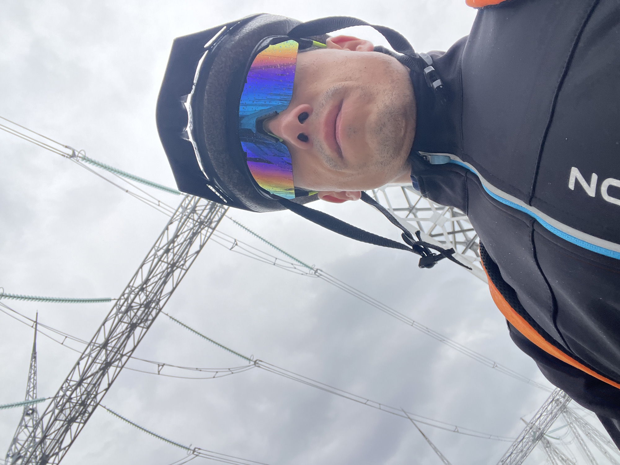 An extreme low-angle selfie captures a person, likely a cyclist, looking upwards against a cloudy sky. The individual wears a black cycling helmet and distinctive rainbow-colored sports sunglasses, which appear to have water droplets on them. Their nose and mouth are visible beneath the glasses. They are dressed in a black cycling jacket with blue, white, and orange accents; the letters "NO" are visible on the right chest area.
The background is dominated by large metal lattice structures, indicative of high-voltage transmission towers or an electrical substation. Numerous power lines with insulators crisscross the grey, overcast sky. The weather appears cloudy, possibly after rain given the droplets on the sunglasses. The setting suggests an industrial or utility area. The image provides a close-up, rotated perspective of the cyclist and their immediate surroundings in Chaikovsky, Russia.